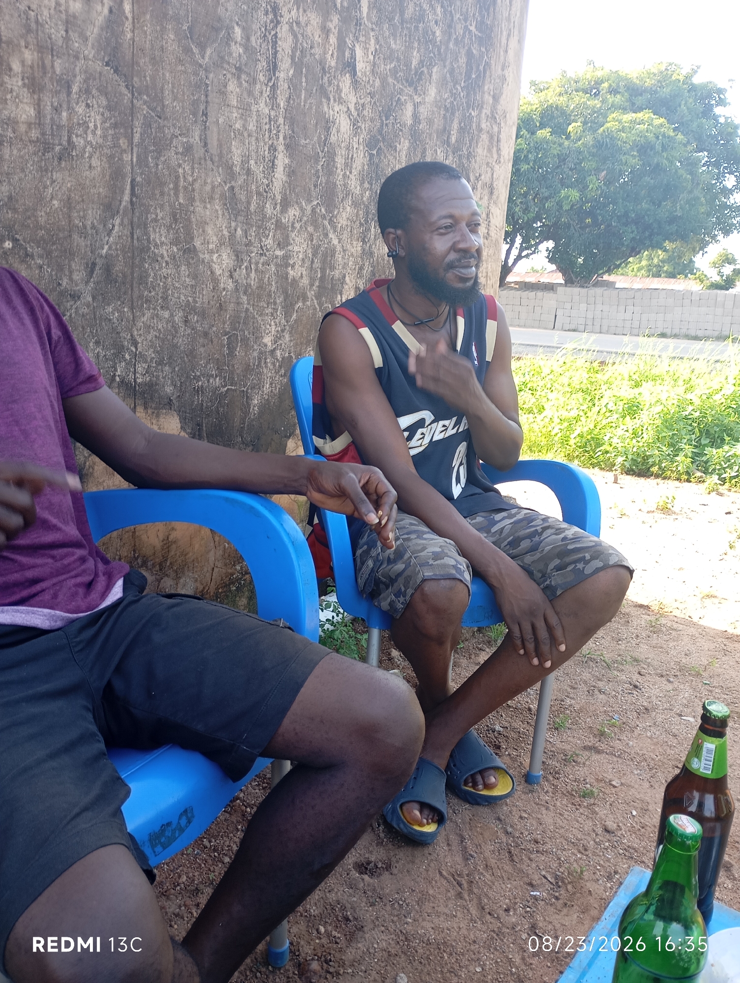 Two men are seated on blue plastic chairs outdoors. The man on the left is wearing a purple t-shirt and black shorts. The man on the right is wearing a sleeveless basketball jersey with the word "LEVEL" visible on it and camouflage shorts. He has a beard and is wearing an earbud. In front of them, on a table, are two bottles of beer, one green and one brown. The background shows a rough, textured wall on the left, and a grassy area with stacked bricks and a tree on the right. The ground is covered in dirt and small stones. The timestamp "08/23/2026 16:35" is visible in the lower right corner, along with "REDMI 13C" in the lower left. The scene appears to be in a casual outdoor setting, possibly in Shendam, Nigeria.