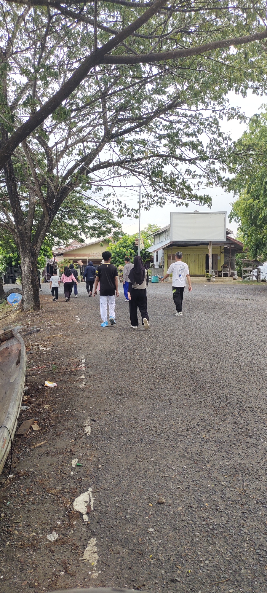 In Lhokseumawe, Indonesia, a group of individuals are walking along a paved road lined with trees and buildings. The scene appears to be in a semi-rural or suburban area, possibly near a residential or recreational facility given the presence of simple structures and open space. The road surface is asphalt with visible gravel and some patches of white marking. To the left, a large tree with extensive branches dominates the foreground, with a discarded wooden boat-like object and debris on the ground beneath it. Further back, several people are walking away from the camera. One person in the foreground wears a black t-shirt and white pants, another wears a black hijab and black pants, and a third wears a white t-shirt with dark pants. Other individuals, including children in lighter clothing, are walking ahead of them. Buildings with light-colored walls and tiled roofs are visible in the background, one of which has a large, blank white screen or billboard mounted on a pole in front of it. The sky is overcast. The overall impression is of a casual outdoor gathering or activity.