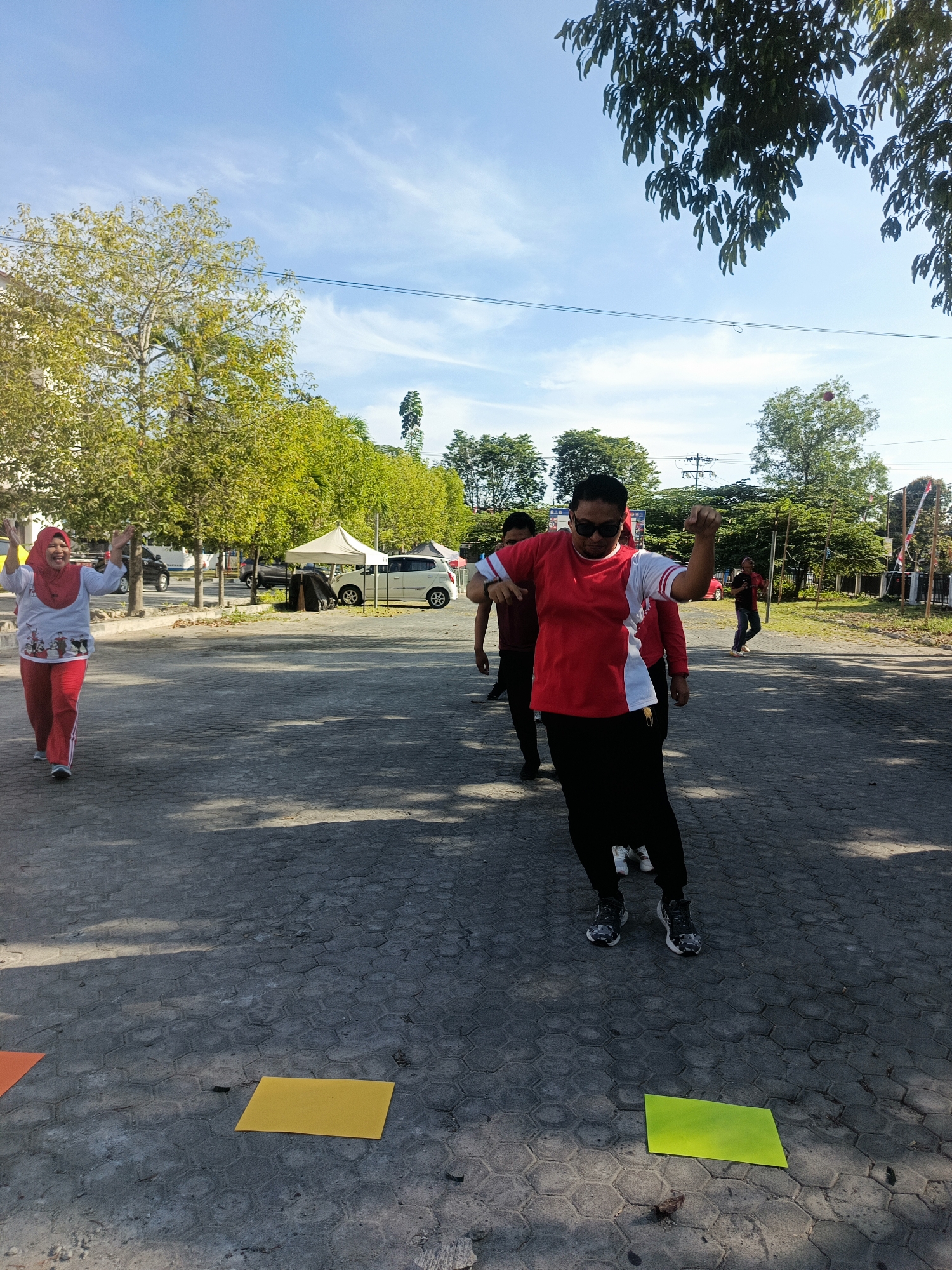 In Gorontalo City, Indonesia, on a paved outdoor area, a group of individuals appears to be engaged in an exercise activity. In the foreground, a person wearing sunglasses, a red and white t-shirt, and black pants is striking a pose with their arms raised. To the left, a woman in a red hijab and red pants is also raising her arms. In the background, other individuals are visible, along with trees, tents, and parked cars. The ground is marked with several colored squares, including orange, yellow, and green, which may indicate a path or markers for the activity. The sky is blue with scattered clouds, and the scene is illuminated by sunlight.