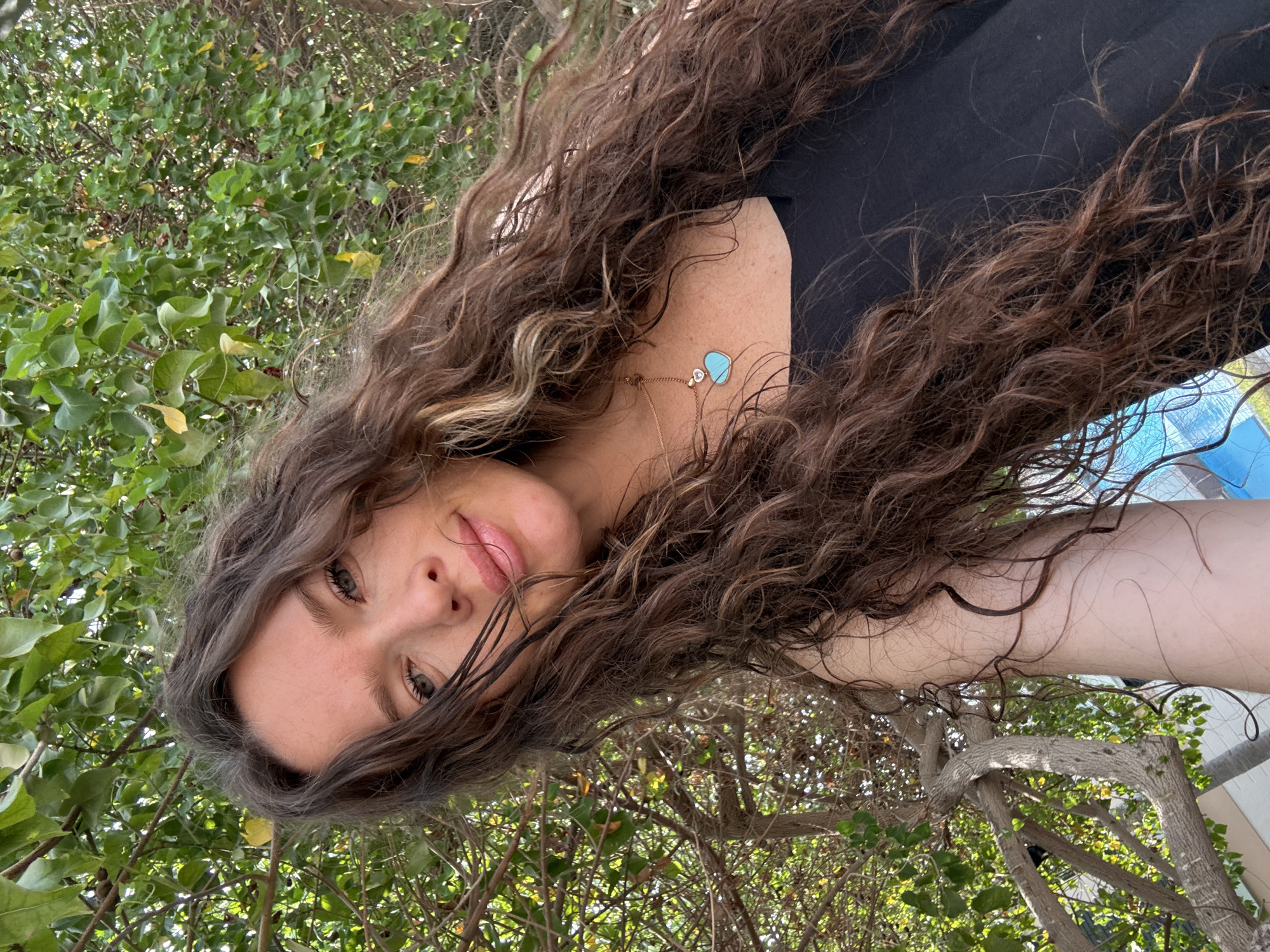 A medium shot captures a woman with long, dark, wavy hair against a backdrop of green foliage and tree branches. She is wearing a black top and a delicate gold necklace with a turquoise heart-shaped pendant. The woman is looking towards the camera with a slight smile. Sunlight filters through the leaves, creating dappled light on her face and hair. Behind her, a blue swimming pool and parts of buildings are visible, suggesting an outdoor setting, potentially a private villa or hotel in Dubai, United Arab Emirates. The image is taken from a slightly low angle, looking up at the woman.