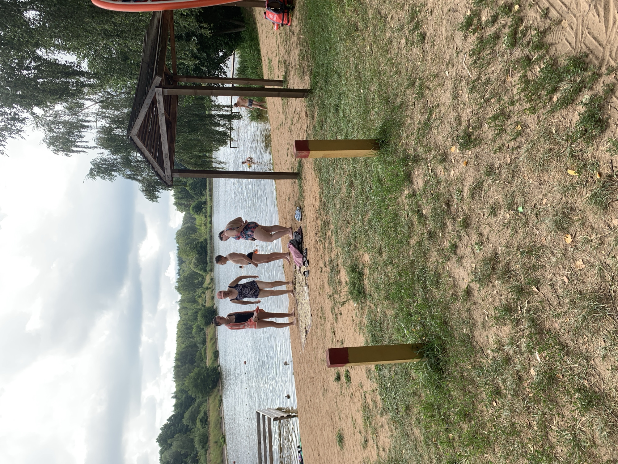 The image depicts a sandy beach area adjacent to a body of water, with a wooded area and a cloudy sky in the background. In the foreground, two wooden posts with red and yellow markings are visible. On the beach, several individuals are present. Four people are standing near the water's edge, three women and one man, all in swimwear. One woman is wearing a pink and orange bikini, another a black and white patterned swimsuit, and the third a patterned blue and pink swimsuit. The man is wearing black swim trunks. A fourth person, a woman wearing a patterned black and white swimsuit, is further to the right. A wooden gazebo structure with a roof is situated on the sand. To the left, a wooden structure extends into the water, and there are several buoys in the water. On the sand near the gazebo, life vests are stacked. Two individuals are in the water: one person is partially submerged, and another person is standing in the water. The environment appears to be a recreational area.