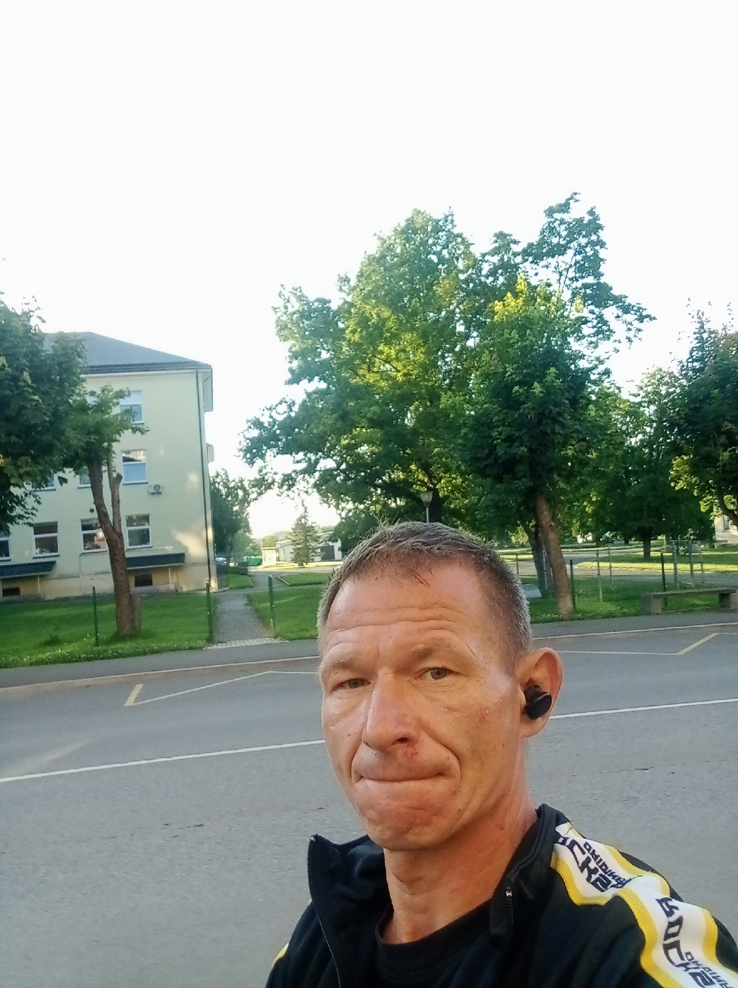 A close-up, selfie-style photograph captures a middle-aged man with short, graying hair and a fair complexion. He is wearing a black zip-up jacket with yellow and white stripes and the word "RICK" repeated in a pattern across the shoulders. A black wireless earbud is visible in his right ear. His mouth is slightly pursed, and a small mark is present on his upper lip. The background is an outdoor urban setting with a paved road and parking lot in the foreground. Behind the road is a sidewalk, a grassy area, and a light-yellow building with multiple windows. Several trees with green foliage are scattered throughout the scene, and a bench is visible in the distance. The sky is bright and overcast, suggesting daytime. The image appears to have been taken in the city of Tapa, Estonia.