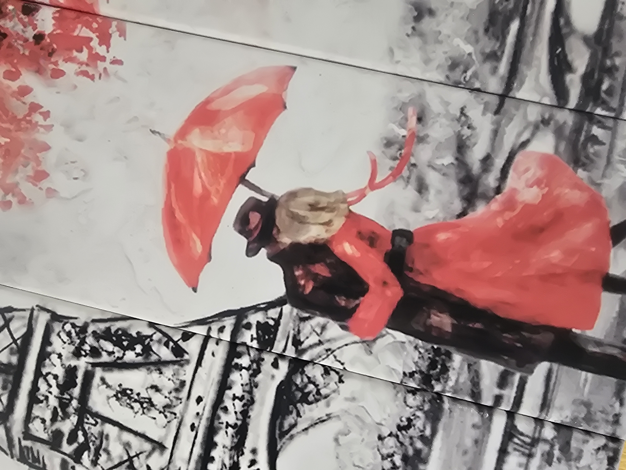 A painting depicts a couple embracing under a red umbrella. The man wears a black coat and hat, while the woman is in a red dress. Behind them, the Eiffel Tower is rendered in black and white. The scene is set against a backdrop of red leaves, suggesting an autumn setting. The artwork is divided into panels, with a visible seam running vertically down the center of the image. The location in Arshan, Russia is not ascertainable from the provided image.