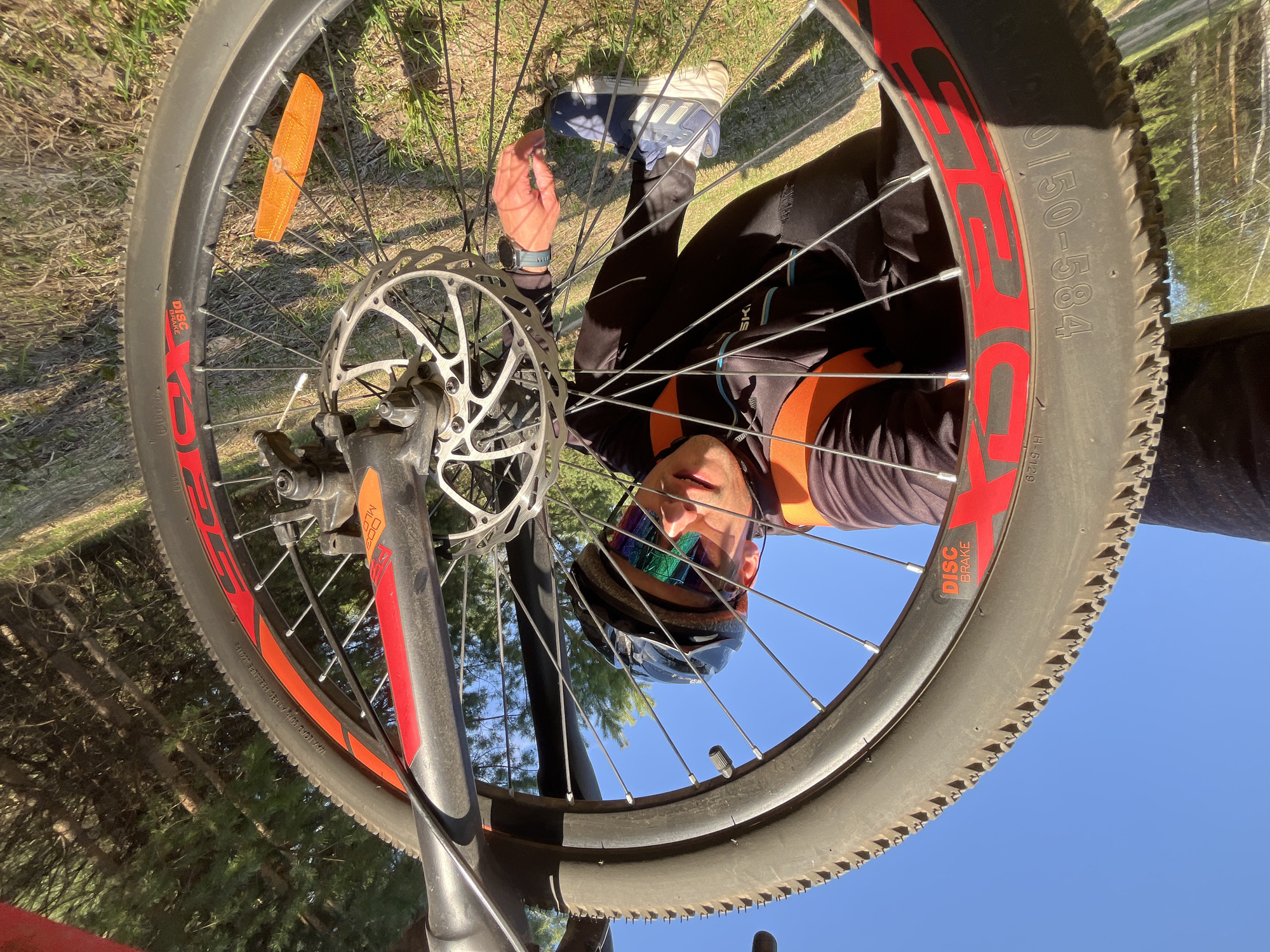 A man is looking through a bicycle wheel. The wheel is in the foreground, and the man's face is in the center of the frame. He is wearing a helmet and sunglasses. The background is a mixture of ground vegetation and trees. The location is unknown, and there is no indication it is in the city of Chaikovsky, Russia.