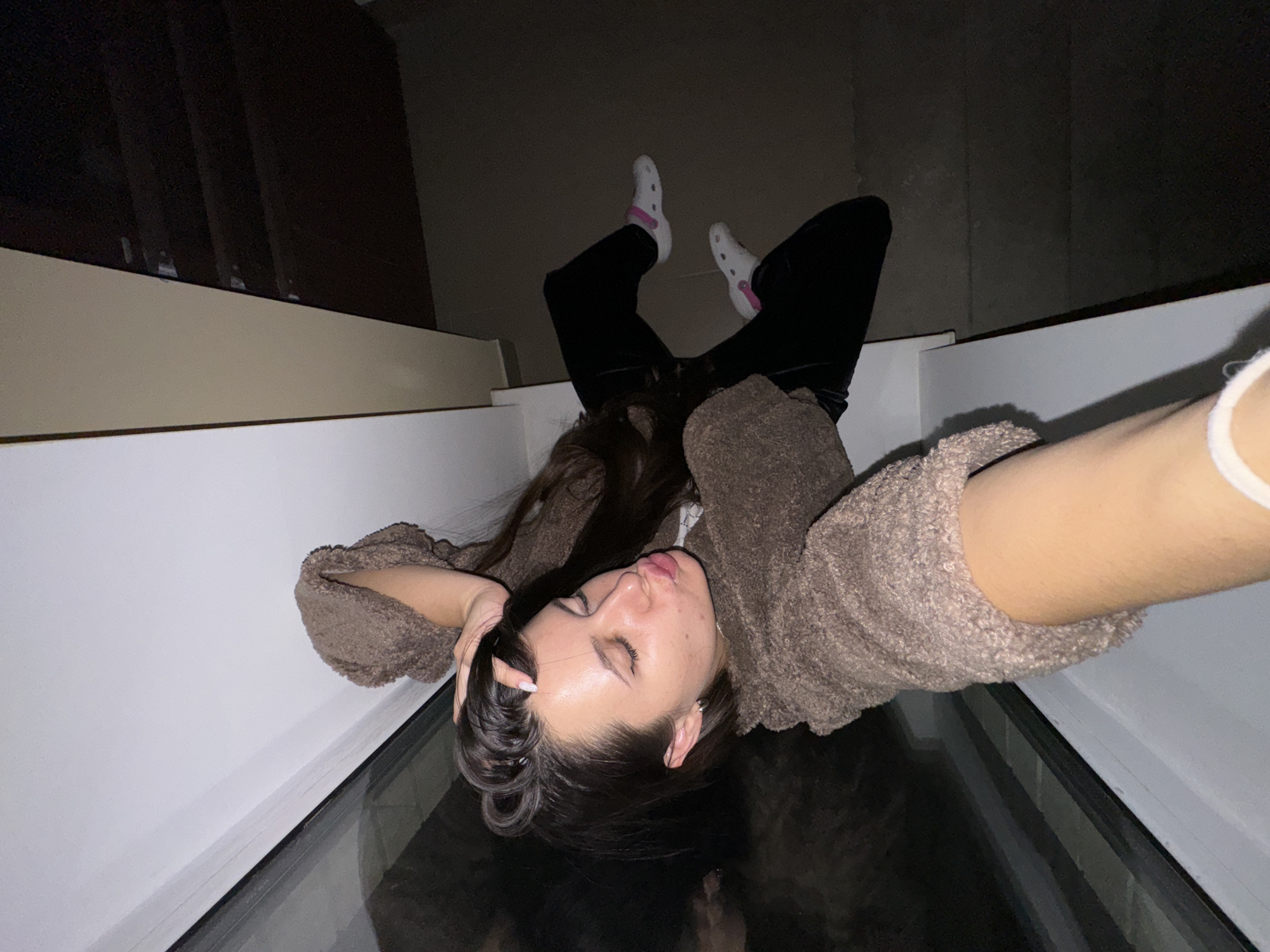 A young woman is lying on a glass surface, likely a skylight or similar architectural feature, within a building. She is wearing black pants and white socks with pink accents, and a brown, textured sweater. The surrounding environment appears to be an interior with beige walls and a dark, possibly wooden structure in the background. The perspective is angled, as if the photograph was taken from directly above, looking down through the glass. It cannot be determined the location in the city of Akulovo (Gp Kubinka), Russia.