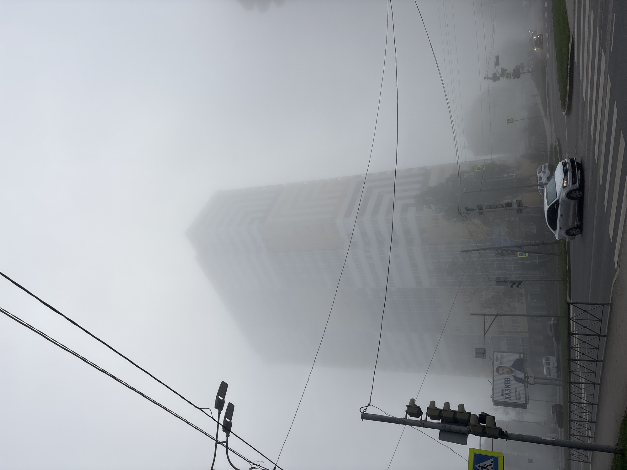 This is a street-level, eye-level photograph taken in the city of Perm, Russia, on a foggy day. A tall, modern apartment building, partially obscured by mist, dominates the background. Its facade features horizontal bands of white and yellow. In the foreground, a street intersection is visible with a crosswalk. A white Volkswagen Polo is crossing the intersection from right to left, with its headlights on. Several other vehicles are present but less distinct due to the fog. Traffic lights are positioned at the intersection, and a pedestrian crossing sign is visible on the left. A large billboard advertising "IREK KHAZIEV" is displayed on the left side of the street. Overhead power lines and streetlights are also present. The overall atmosphere is somber due to the dense fog, which significantly reduces visibility.