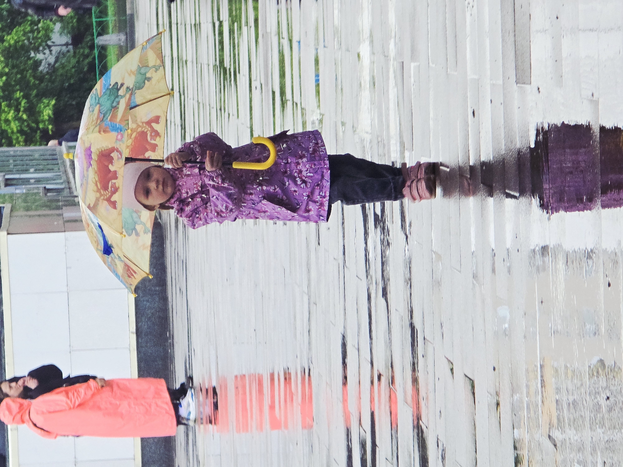 A child stands under a dinosaur-themed umbrella in what appears to be a rainy urban setting. The child, dressed in a purple floral jacket and dark pants, is holding the umbrella with both hands. The umbrella's design includes cartoon dinosaurs in various colors.
The ground is wet, creating reflections of the people and objects in the scene. The surface appears to be composed of light-colored rectangular paving stones. In the lower left corner, a person in a coral-colored jacket is visible.
Based on the surroundings and context, the location may be in Moscow, Russia, although no specific landmarks confirming this are visible. The scene suggests a moment of daily life during inclement weather.