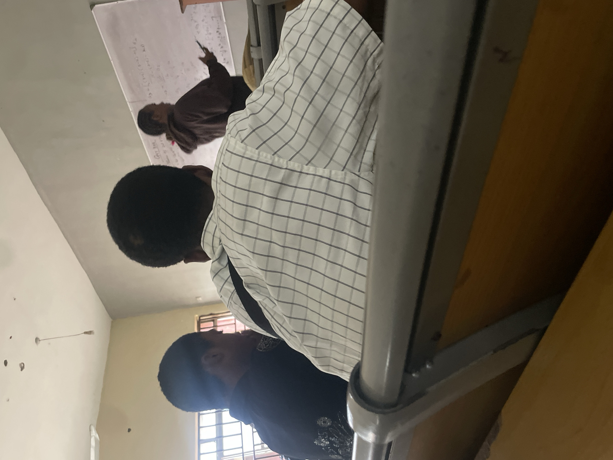 This image depicts an indoor scene, likely a classroom, in Ihiagwa, Nigeria. In the foreground, two young men are seated in what appear to be tiered benches, facing away from the camera. The man on the right is wearing a white and black checkered shirt, while the man on the left is wearing a dark shirt with metallic embellishments on the back.
In the background, a teacher, possibly male, is standing near a whiteboard and gesturing with a stylus or marker in his right hand, as if explaining something written on the board. The whiteboard contains handwritten mathematical equations and text in English. The room has a simple ceiling with a hanging light fixture and windows with blinds on the left side of the frame. The overall environment suggests an educational setting.
