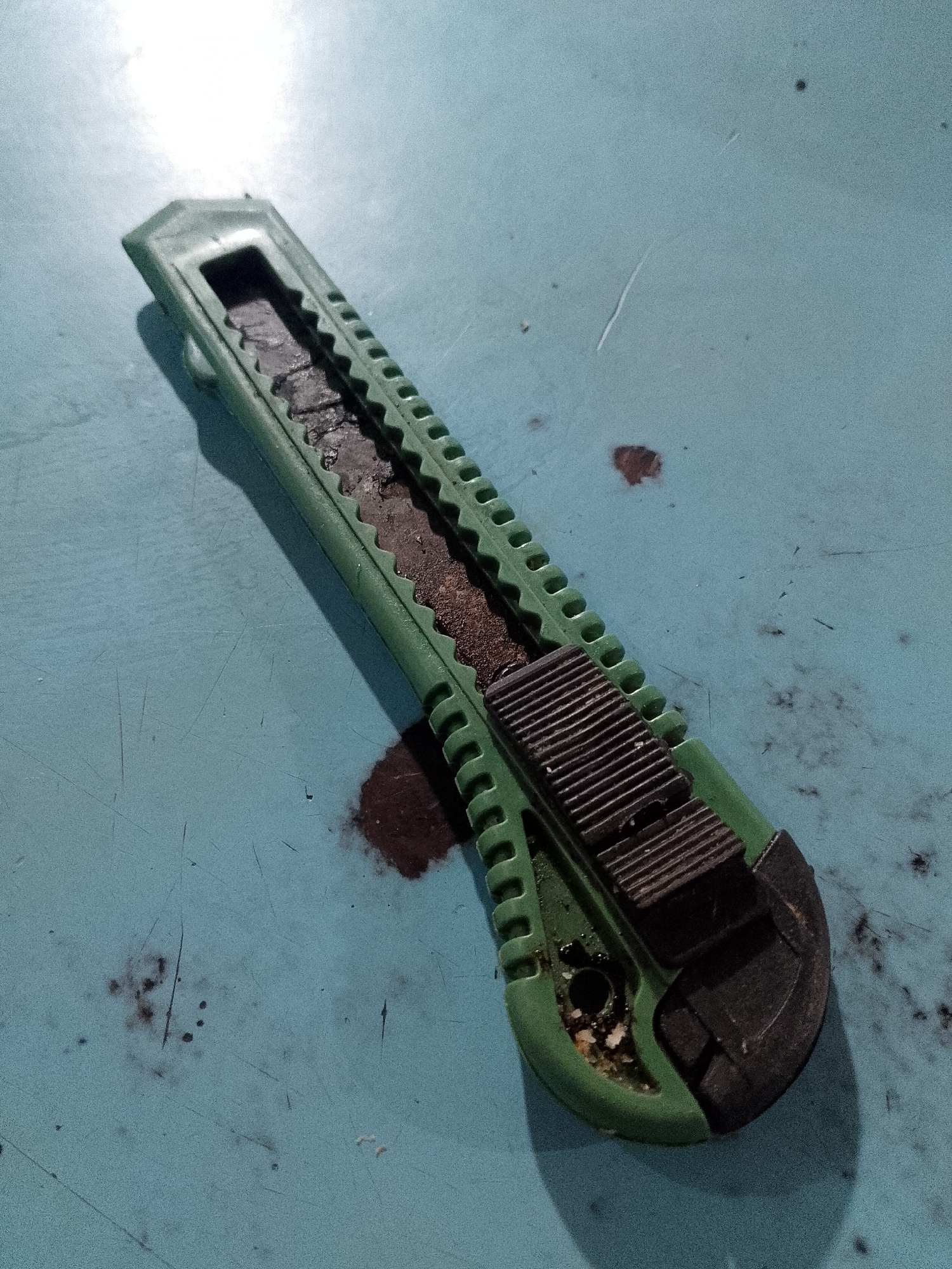 The image shows a green and black utility knife lying on a light blue surface. The knife appears worn and contains a dark substance in the blade channel. There are also dark stains on the light blue surface around the knife. No people, subjects, actions, or interactions are visible. The location is stated to be Lhokseumawe, Indonesia, though this is not visually verifiable from the image itself.