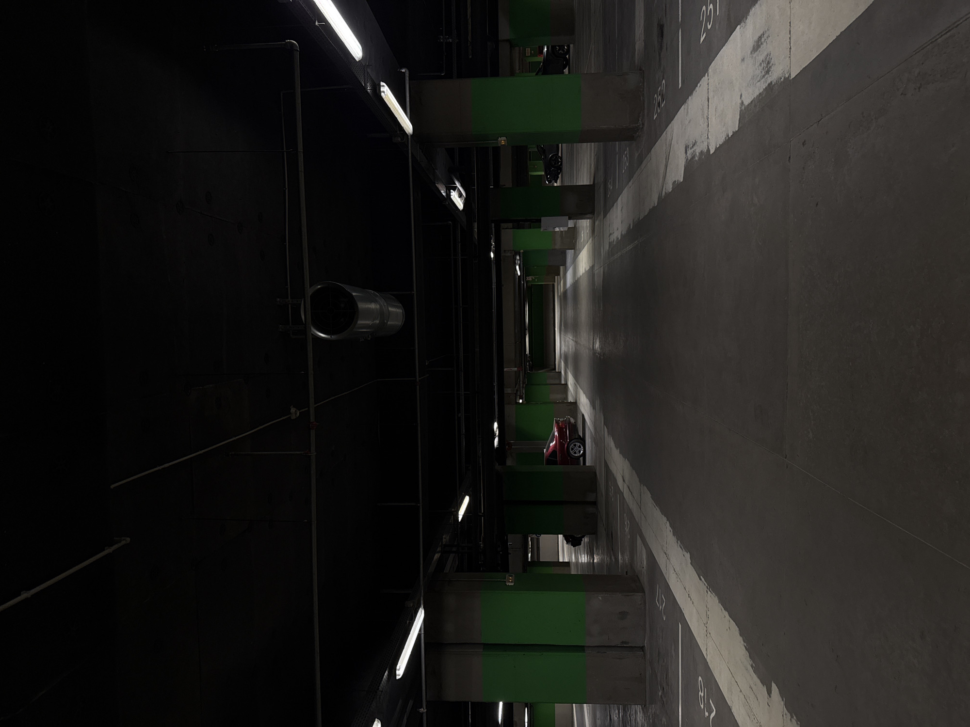 The image shows a dimly lit, underground parking garage. Concrete columns painted with green stripes support the structure. Fluorescent lights are visible on the ceiling. On the right, a gray wall is marked with parking space numbers in white. In the distance, a red car is parked. A cylindrical ventilation duct is visible on the left. It is unknown if this photo was taken in Sofia, Bulgaria.
