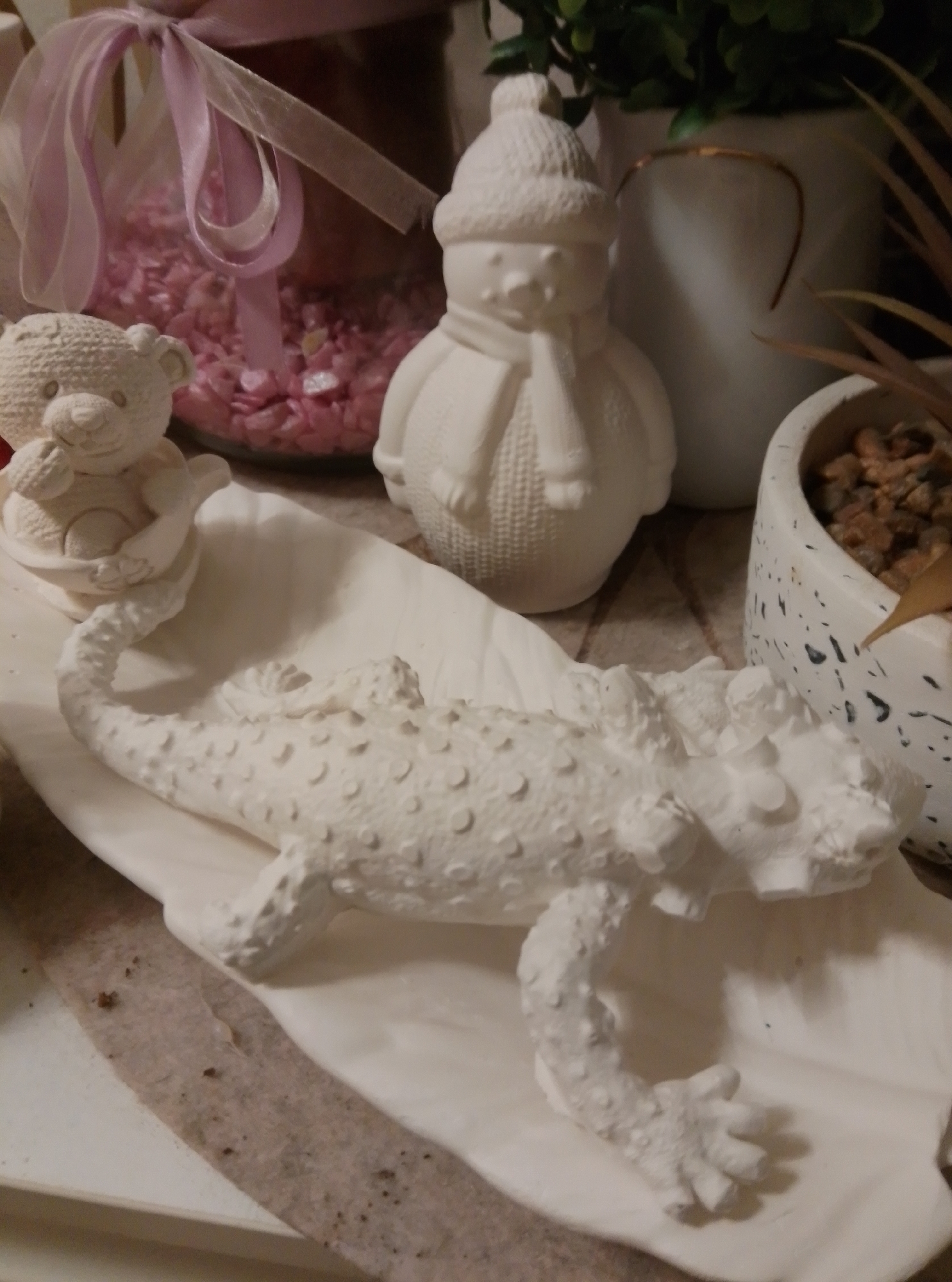 The image shows a collection of ceramic figurines and plants on a table. In the foreground, a white ceramic lizard is lying on its side on a large, white, shell-shaped platform. The lizard has a textured body with raised bumps and its limbs are bent. To the left of the lizard, a small white ceramic teddy bear is sitting in a small white bowl, with one paw raised. Behind the figurines, there is a glass jar filled with pink decorative stones, tied with a pink and white ribbon. Further back, a white ceramic snowman figurine with a scarf and hat stands next to a potted green plant in a white speckled pot. The overall setting appears to be a tabletop display, possibly in a home or a shop. The image was taken in the city of Wedel, Germany.