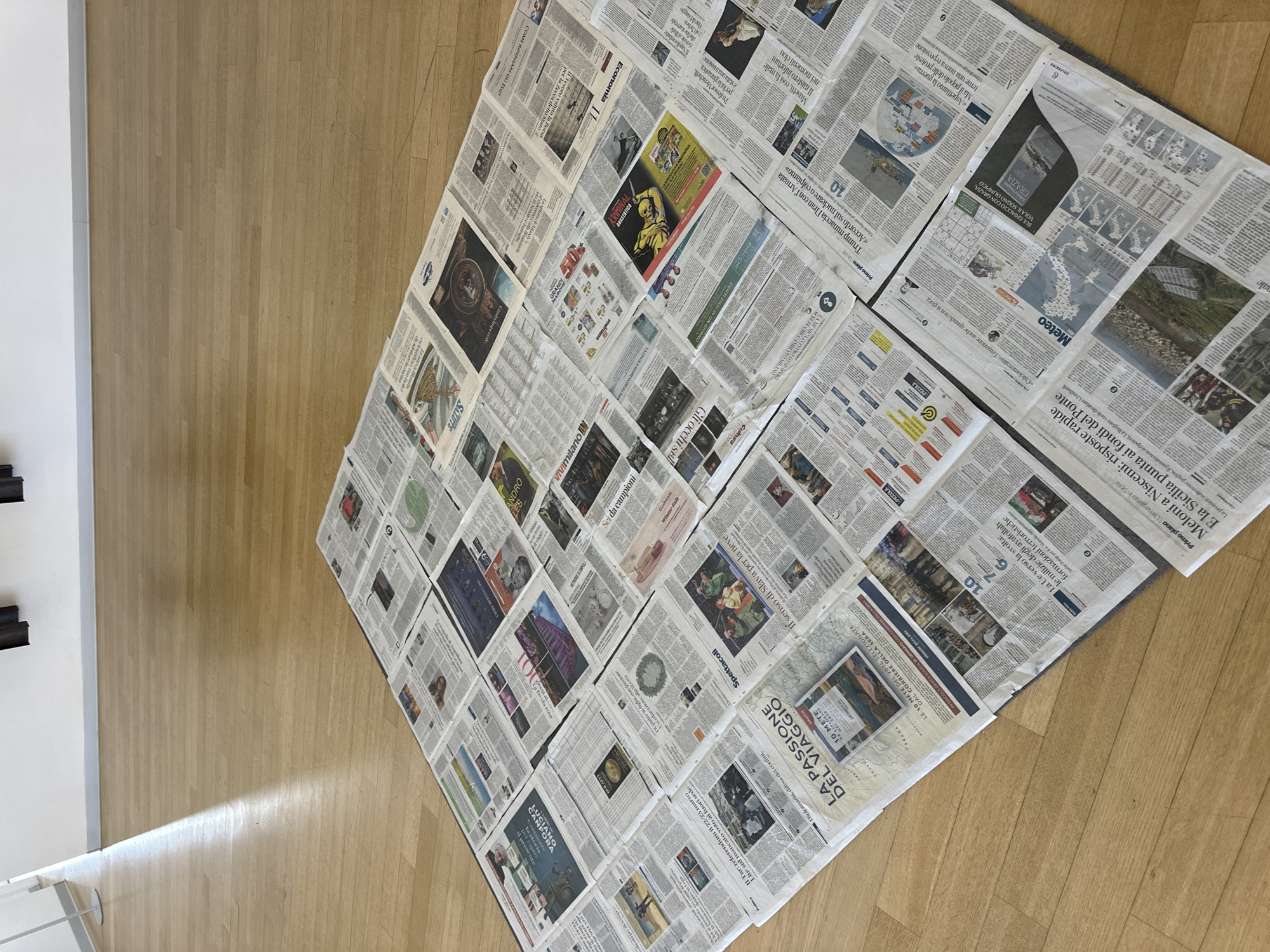 The scene shows a floor covered with numerous newspaper pages. The newspapers are spread out in a roughly diamond shape on a light-colored wooden floor. The newsprint displays a mix of text, headlines, and images, some in color and some in black and white. The articles are in Italian. Prominent words and sections visible include "MILANO," "LA PASSIONE DEL VIAGGIO," "GRATIS," "Meteo," and names like "Meloni" and "Trump." Part of a white wall with a dark-colored decoration on it is visible to the left side of the frame. Based on the "MILANO" headline, it is in the city of Milan, Italy.