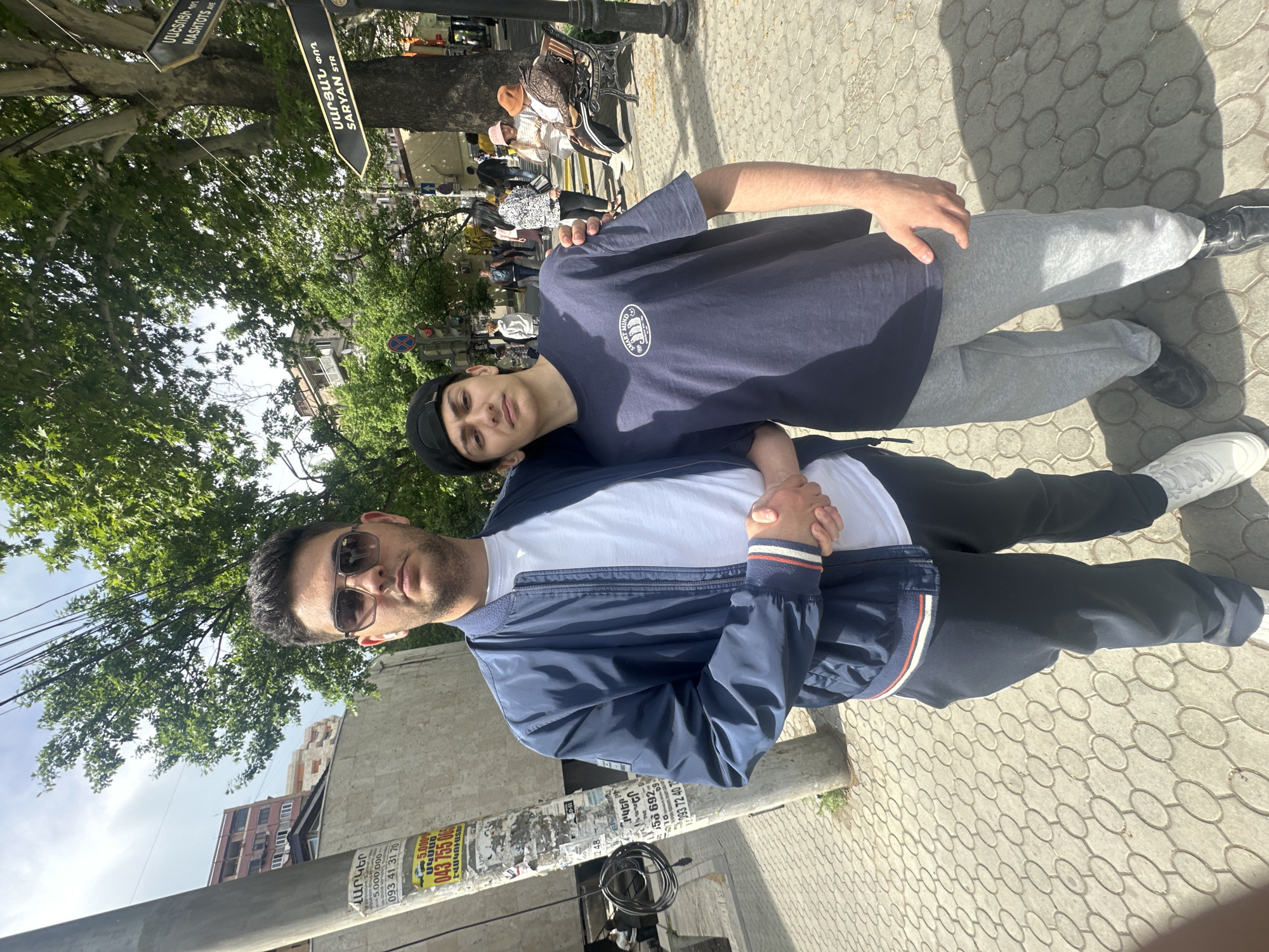 Two young men are standing close to each other on a paved area. The man on the right is wearing a navy t-shirt, gray sweatpants, and a black beanie. The man on the left is wearing sunglasses, a white t-shirt, and a navy jacket with white and red stripes on the cuffs. They are holding hands. Behind them is a street with people, trees, and a street sign that says "SARYAN STR" and "MASHTOTS AVE" in Armenian and English, indicating they are in Yerevan, Armenia. There is also a lamp post with advertisements on it.