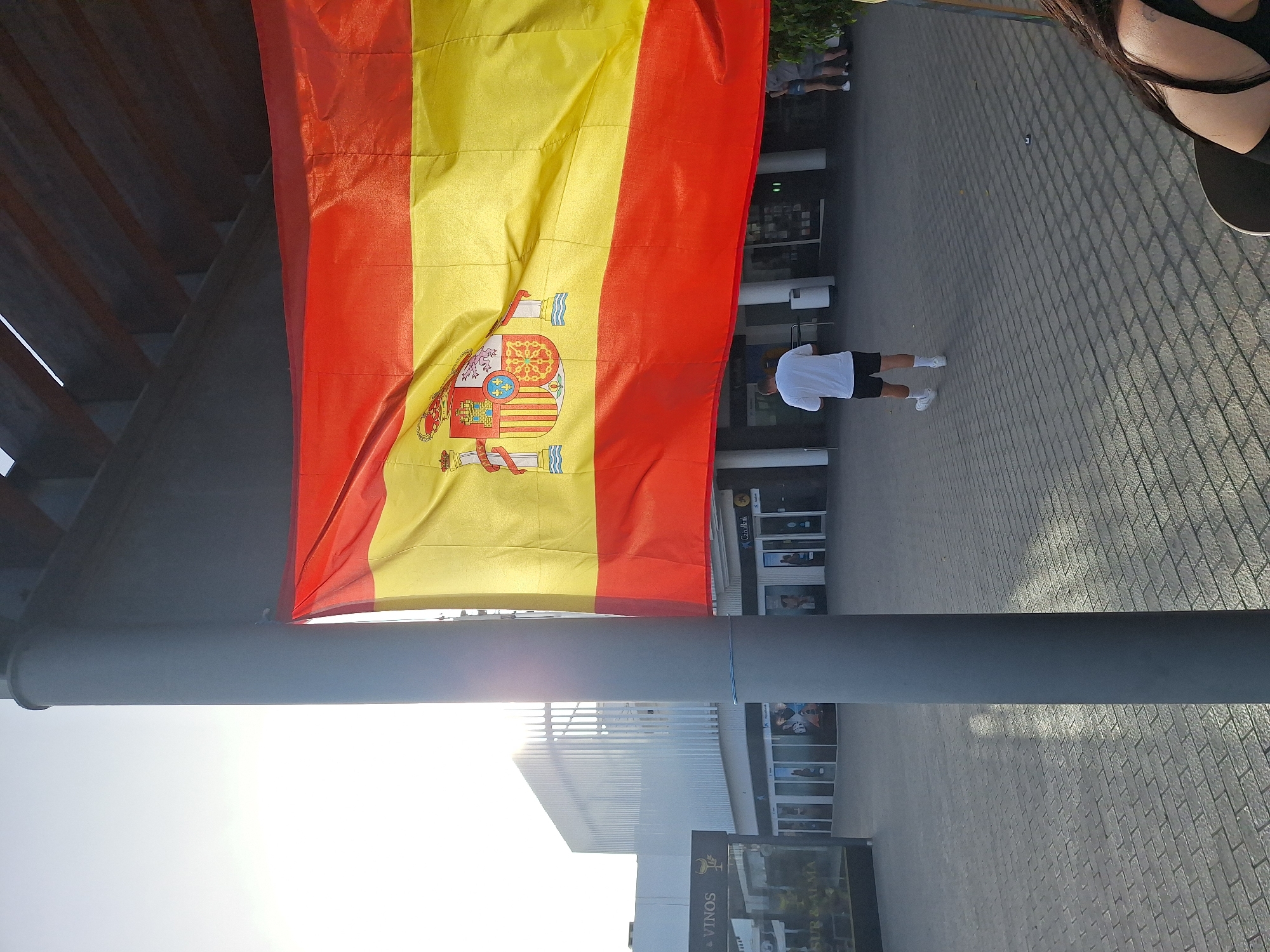 The image captures a sunny outdoor scene, likely in the city of San Bartolomé, Spain, given the Spanish flag prominently displayed. The flag, featuring red and yellow horizontal stripes with the Spanish coat of arms, is draped over a large, grey pole, creating a central visual element. In the background, a modern white building with vertical louvers is visible, and several storefronts, including one labeled "CaixaBank," line a paved plaza. A few people are present: a man in a white t-shirt and black shorts is walking away from the camera, and a couple stands near the storefronts on the right. The ground is paved with rectangular grey bricks, and sunlight casts dappled shadows across the scene. A portion of a woman's shoulder and back is visible in the lower right corner of the frame, suggesting she is seated. The overall atmosphere appears to be that of a public space, possibly a commercial area or promenade, on a bright day.