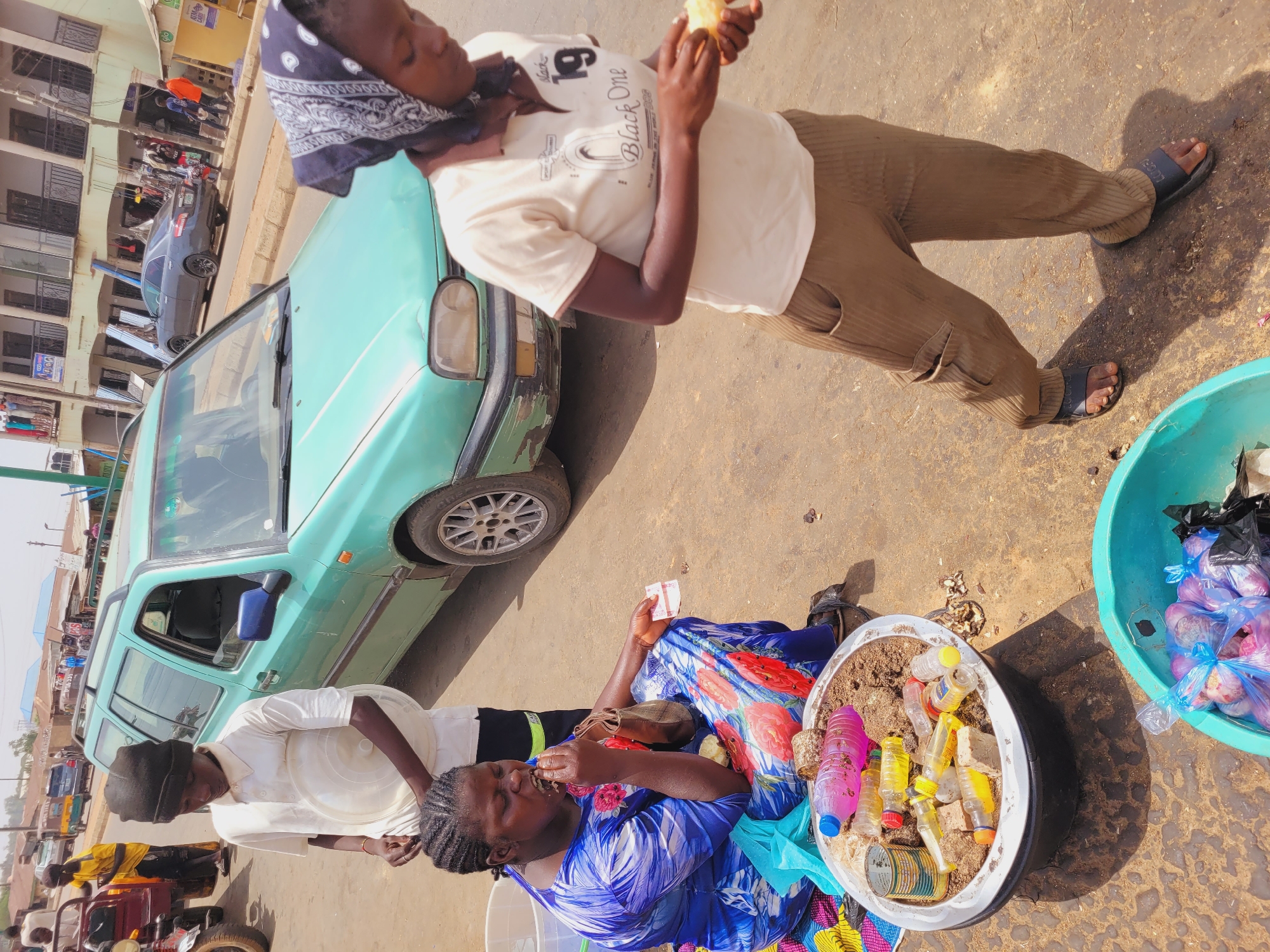 The image depicts a scene in Bukuru, Nigeria, on a sunny day. Two individuals are seated next to a green car and an assortment of goods for sale. One woman, wearing a blue and red patterned dress, is eating while holding money in her hand. Next to her is a white basin filled with water bottles and other small containers. Another person, wearing a white shirt, black pants, and sandals, is standing, also holding food to their mouth. The car is parked on a paved area, and behind the scene, buildings and other vehicles, including a motorcycle, are visible, indicating an urban setting. Another woman wearing a head scarf stands adjacent to the vehicle, examining what appears to be a potato.