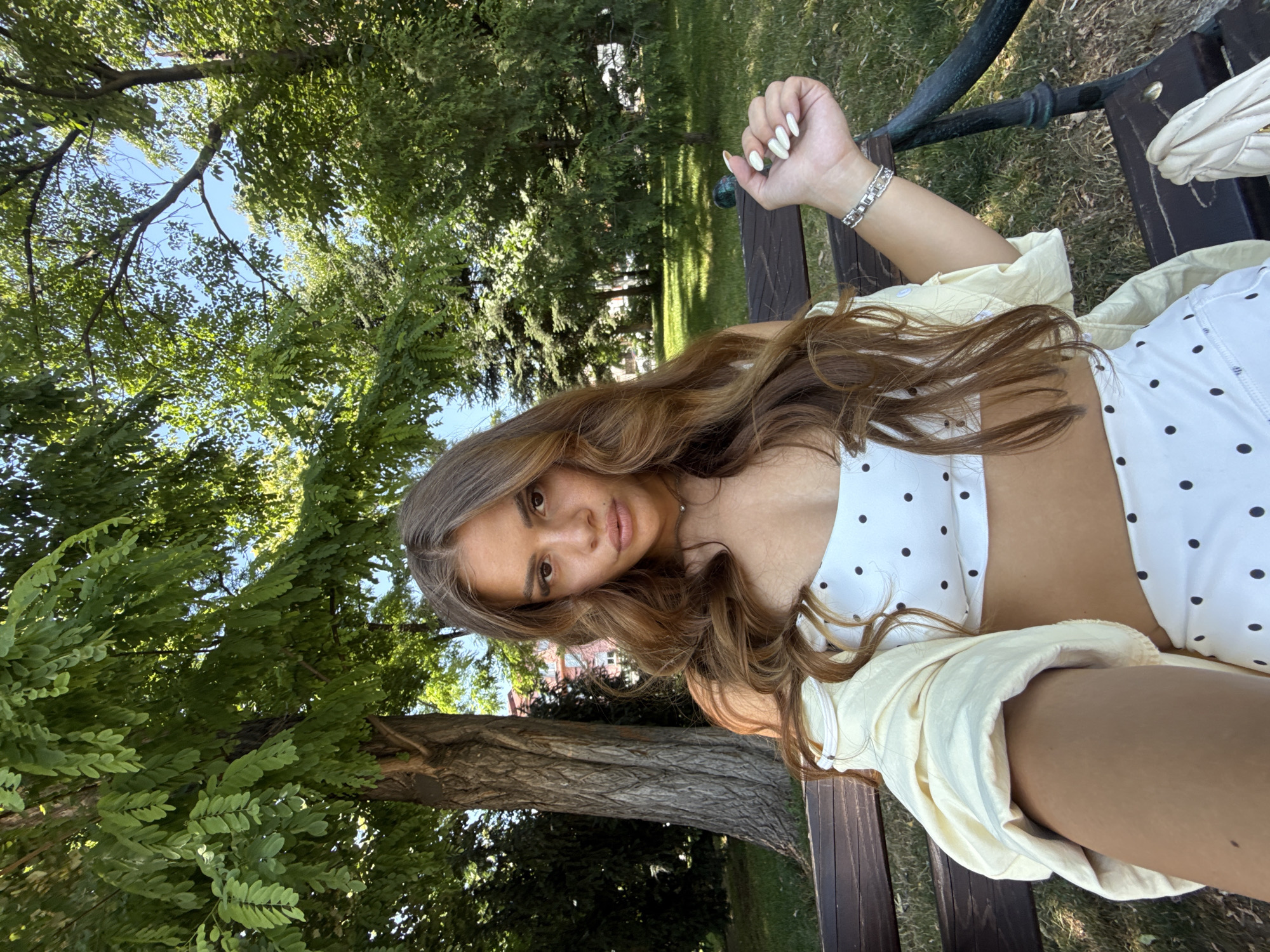 The image shows a young woman with long, brown, wavy hair sitting on a park bench. She is wearing a white crop top with black polka dots and a light yellow unbuttoned shirt over it. She is wearing a silver bracelet on her left wrist and has white nail polish on her fingernails. She is looking towards the camera with a relaxed expression. The background is a park with lush green trees and grass, indicating it is likely daytime and the weather is pleasant. The bench appears to be made of dark wood. In the background, there are more trees and the hint of buildings, suggesting an urban park setting. The overall mood is casual and summery. The image was taken in Plovdiv, Bulgaria.