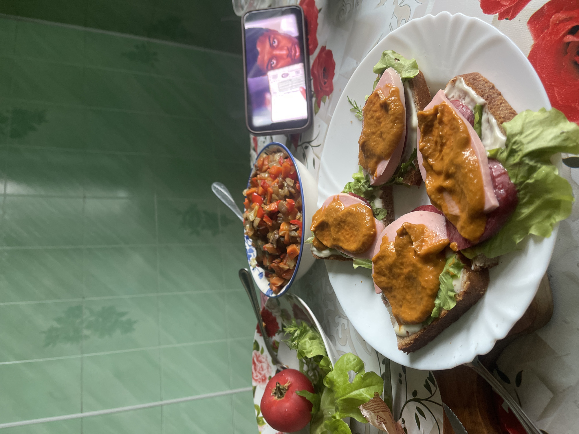 In an indoor setting, possibly a kitchen or dining area, a table is laid with a floral-patterned tablecloth. On the table, there are several open-faced sandwiches on a white plate. Each sandwich consists of a slice of dark bread topped with lettuce, a slice of pink ham or sausage, and a dollop of orange sauce. To the left of the sandwiches, a shallow bowl contains a mixture of chopped vegetables, likely a salad or stew. Adjacent to the vegetable bowl is a white plate with a single red tomato and lettuce leaves. In the background, a wall is covered with light green tiled squares. A mobile phone, placed on the table, displays a paused video or image of a person's face. The lighting suggests an indoor environment during the daytime. The scene does not provide specific visual cues to pinpoint the location within the city of Красное, Russia.