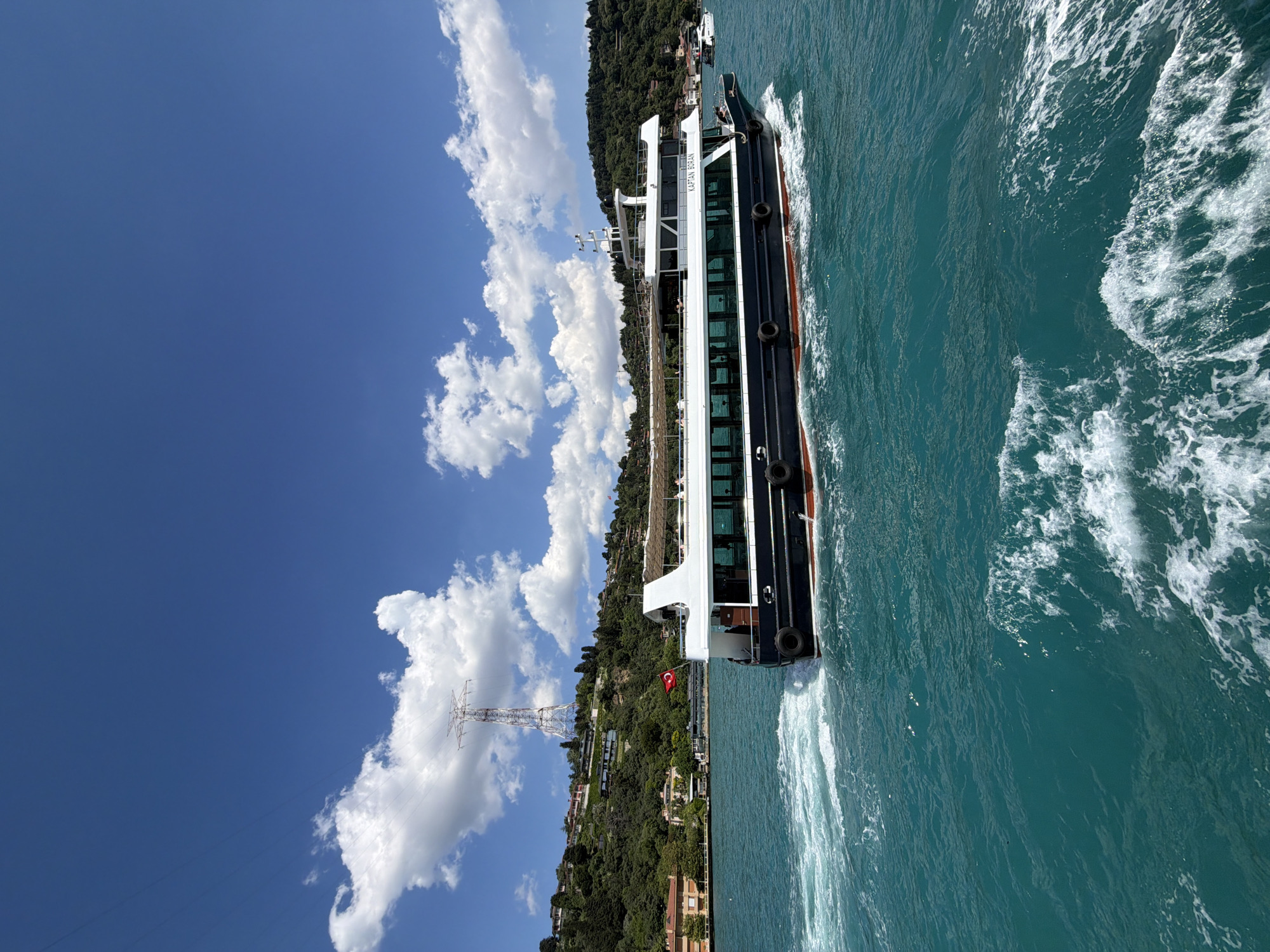 A ferry, named "KAPTAN BORAN," travels through the turquoise waters of the Bosphorus Strait. The ferry has a dark blue hull with a white upper deck and windows. The boat is leaving a white wake behind it. On the left side of the image, a lush, green hillside is dotted with houses. A large, prominent electrical tower stands tall on the hillside under a bright blue sky filled with fluffy white clouds. A Turkish flag is visible on the ferry.
