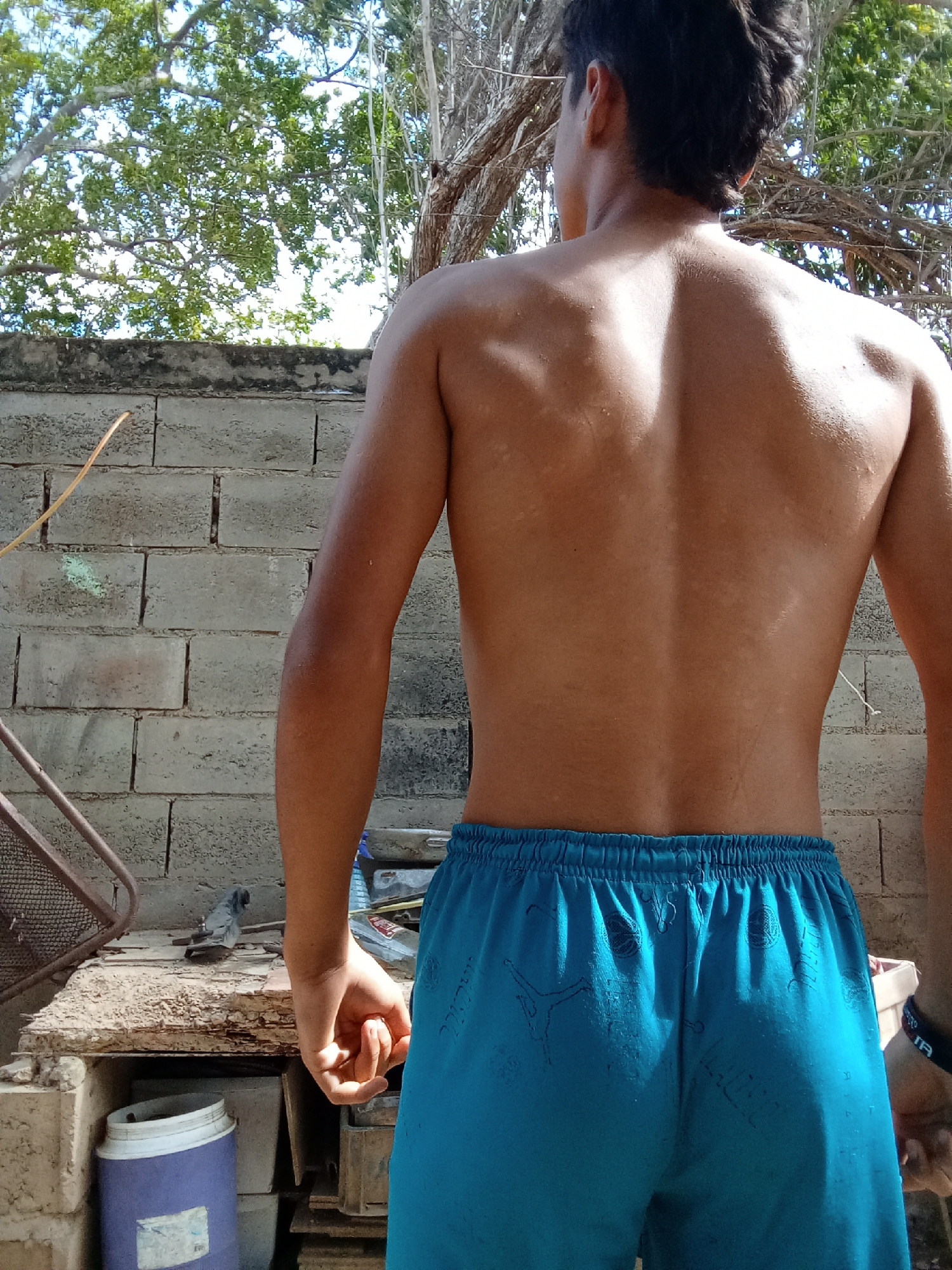 A medium shot shows a shirtless man with tan skin and short dark hair, standing outside. The man faces away from the viewer, revealing his back. He is wearing blue shorts with images on the fabric. The man is standing in front of a rough, light gray brick wall. To the left of the man, there's a partially visible wooden table with assorted items, including a blue and white bucket. Behind the man, there are trees with dense green foliage and bare branches. Based on the location information, the scene is likely in the city of Juan Griego, Venezuela.
