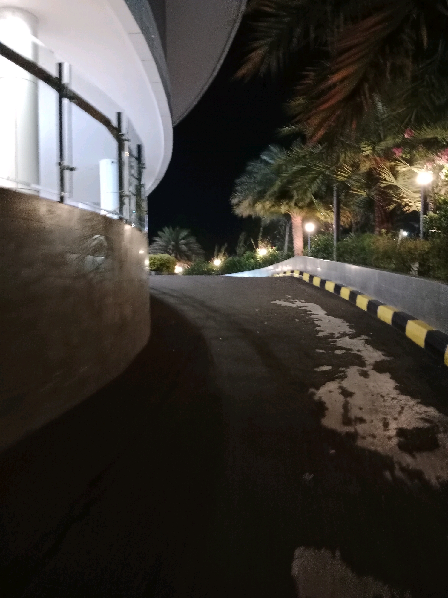 The image shows a dimly lit outdoor scene at night, likely a pathway or driveway with palm trees and some vegetation. A modern building with a curved white facade is visible on the left. The pathway is paved and has a wet appearance in places, reflecting the light from nearby lamps. A black and yellow striped barrier lines the right side of the pathway, separating it from a raised area with vegetation. Streetlights or landscape lighting are visible amongst the trees, casting a warm glow. The overall impression is one of an exterior area at a commercial or residential complex at night. There is no information in the image that would place it in Cianjur, Indonesia.