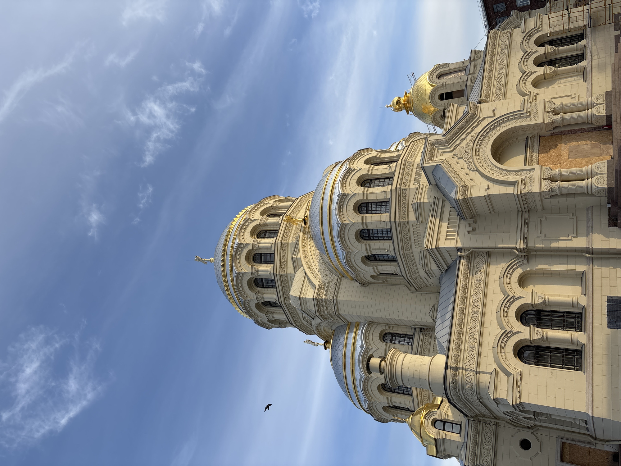 The image shows a low-angle view of the Naval Cathedral of Saint Nicholas in Kronstadt, Saint Petersburg, Russia. The cathedral is a large, ornate building with a pale exterior and gold and silver domes. The domes are layered, decreasing in size as they ascend, and feature decorative elements like crosses and arched windows. The building's walls exhibit intricate carvings and moldings. A solitary bird is visible against the blue sky with wispy clouds. Construction or restoration work is evident on parts of the building, with scaffolding and temporary coverings present.