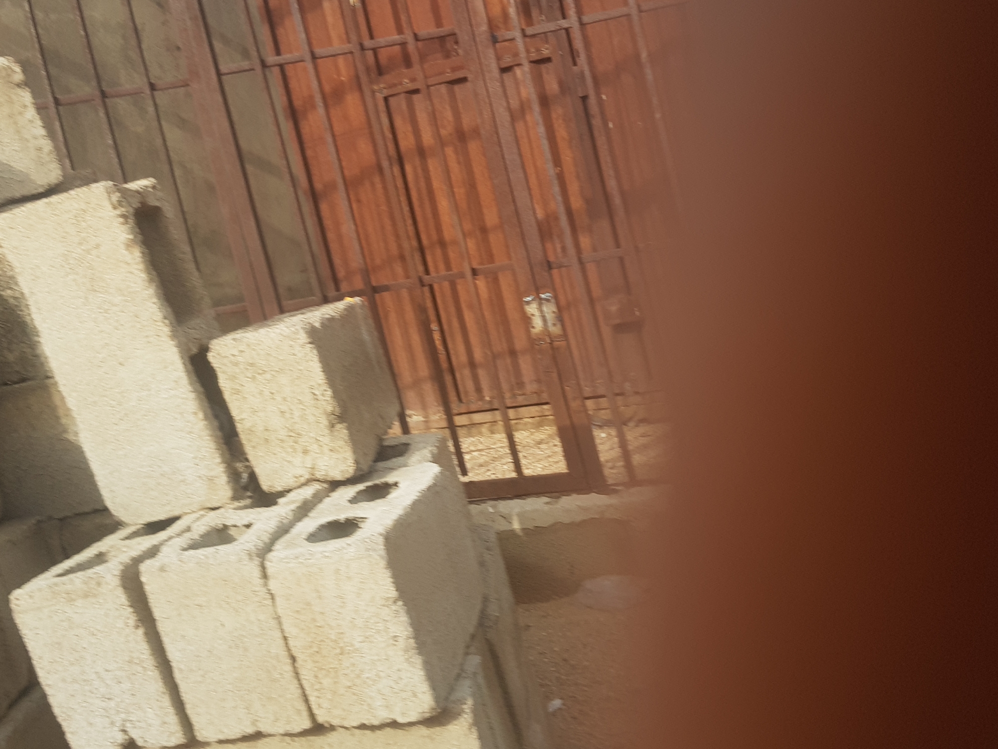 The image shows a stack of gray cinder blocks in front of a reddish-brown metal gate or door. The ground is sandy. The location is possibly in Dabawa, Nigeria. A blurry object obscures the right side of the image.