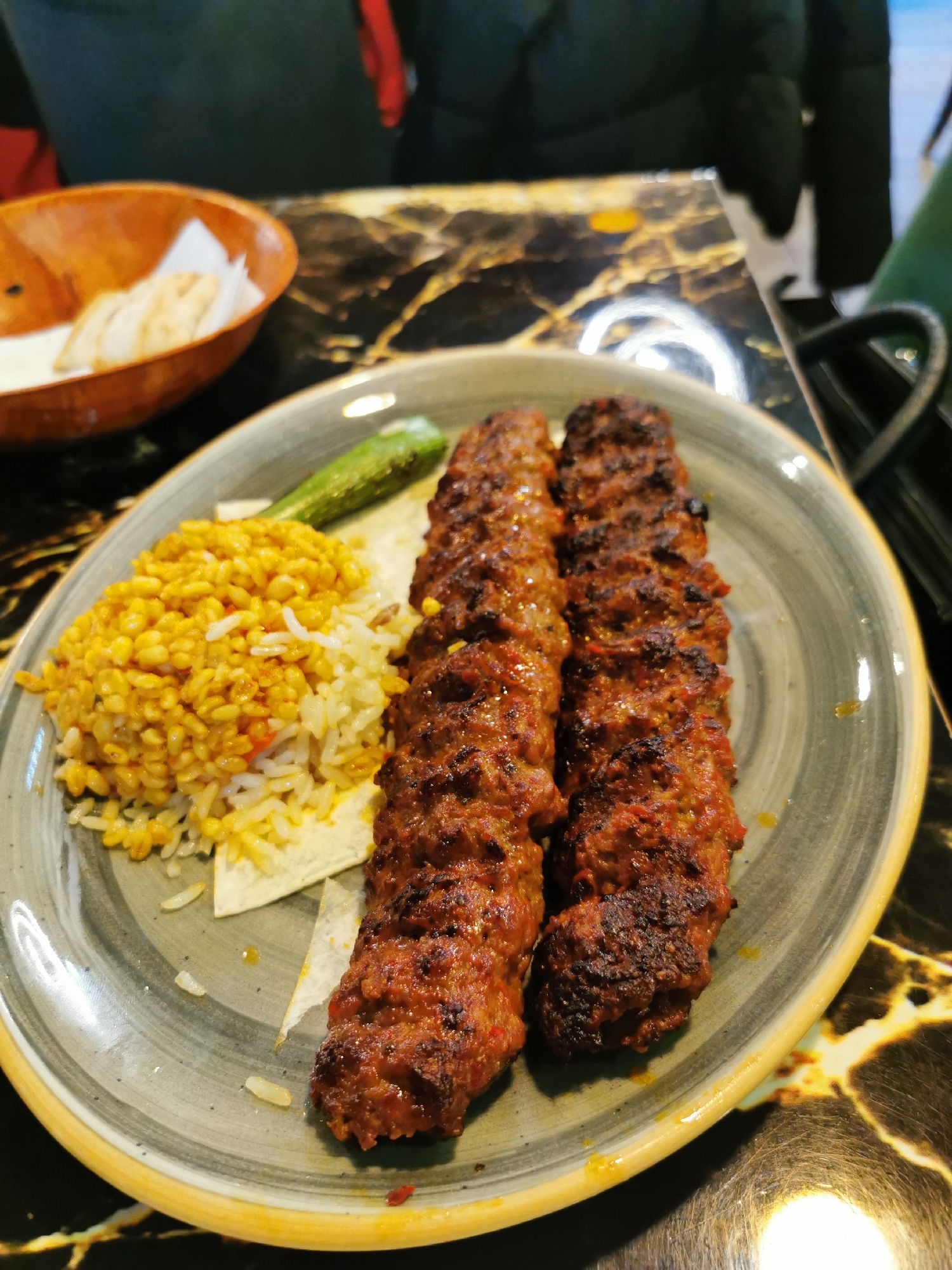 On a table with a dark marble-like surface, a plate holds two skewers of grilled meat, a mound of yellow rice and bulgur, a green chili pepper, and flatbread pieces. A wooden bowl with bread is visible in the upper left. In the background, people in dark clothing are present. The location is inferred from the image details, it is a restaurant in London, United Kingdom.