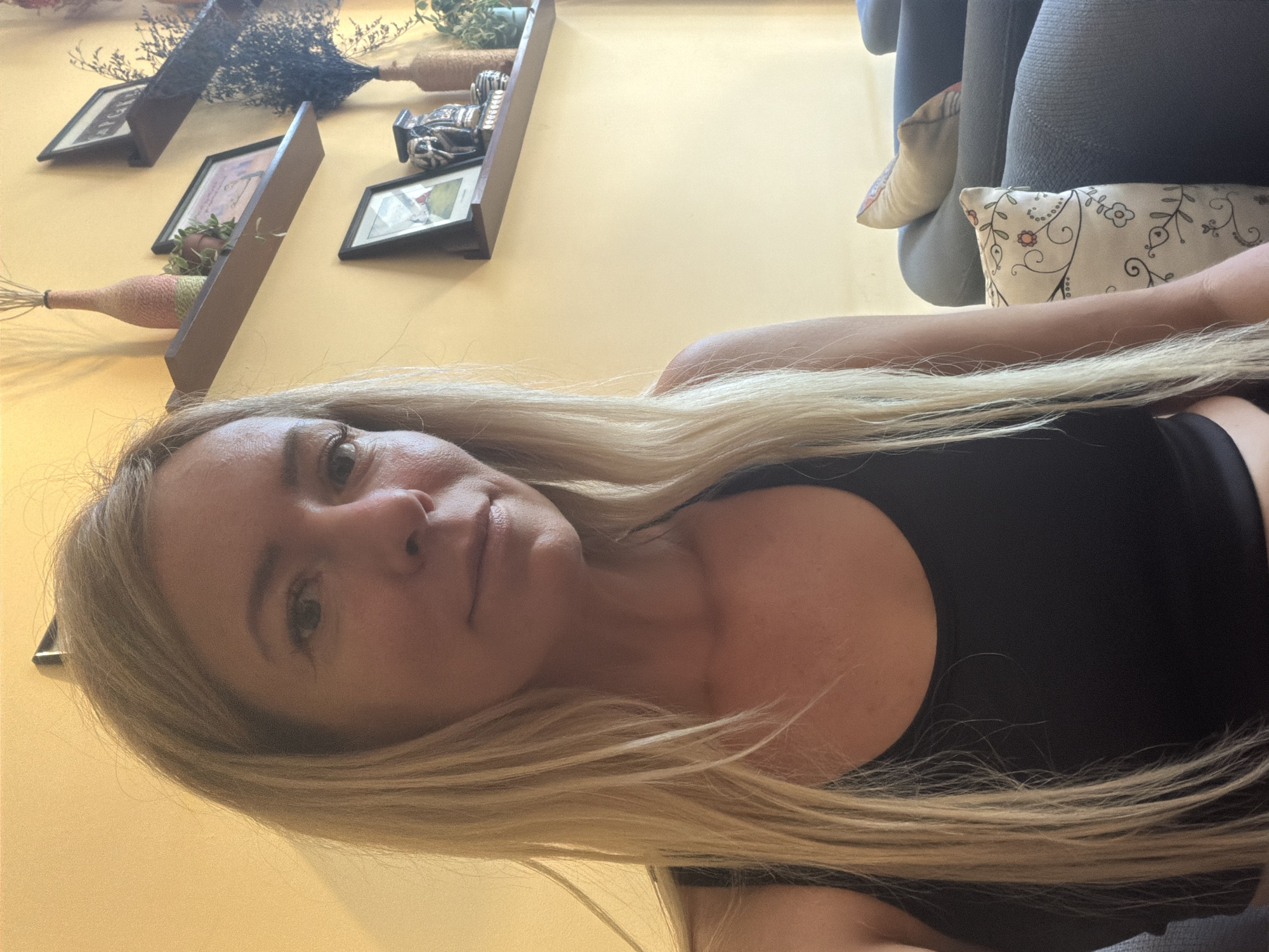 A woman with long blonde hair, wearing a black tank top, is shown from the chest up in a selfie. She is seated in a room with yellow walls. On the wall behind her, there are several shelves displaying decorative items, including framed pictures, vases, and dried plants. To the right of the woman, a dark blue couch with patterned pillows is visible. The environment suggests an indoor setting, possibly a cafe or lounge area, in Cam Ly, Đà Lạt, Vietnam.
