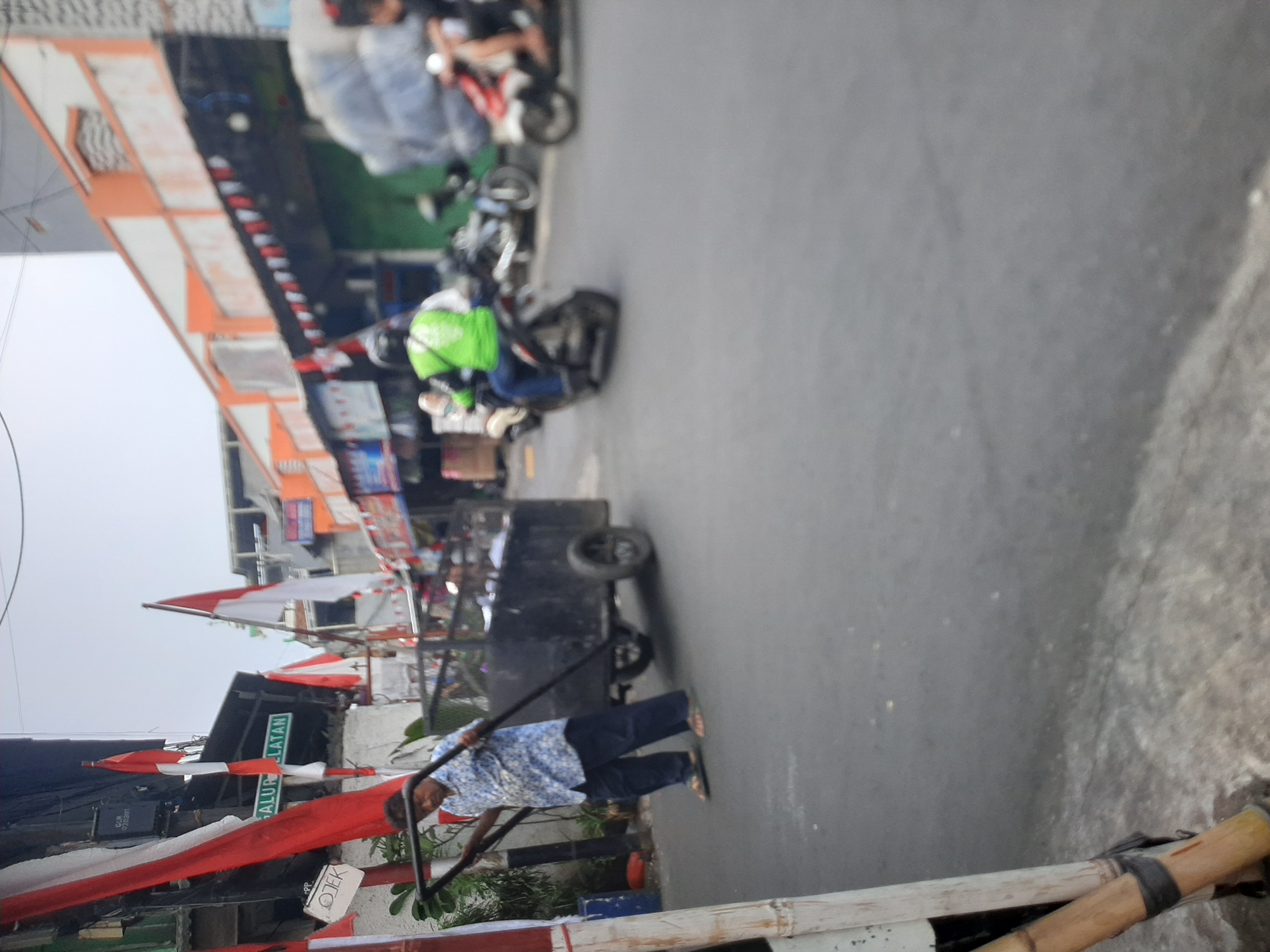 The image captures a street scene in Central Jakarta, Indonesia, likely on a cloudy day. A man in a patterned blue shirt and dark pants is pushing a large, dark-colored cart with two wheels down the asphalt road. The cart appears to be filled with various items, partially covered. To the left, a pole with red and white Indonesian flags is visible, along with a sign that reads "OJEK". In the background, several motorcycles are on the road. One motorcycle has a rider wearing a bright green jacket, and another shows a rider in red and white. Buildings with orange and white facades line the street, adorned with more red and white flags, suggesting a patriotic or celebratory atmosphere. The overall impression is one of everyday activity in an urban environment.