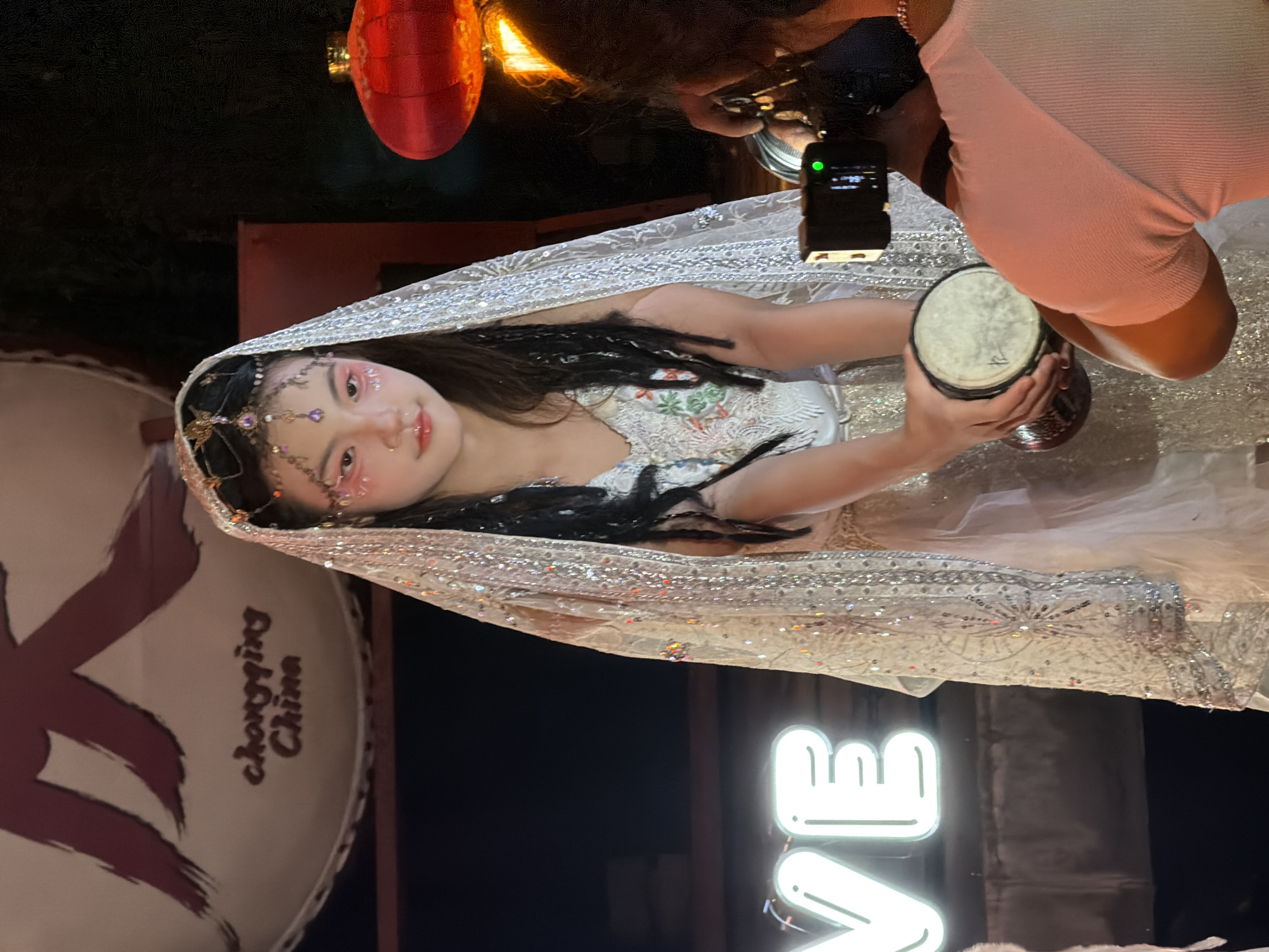 In Yuzhong District, China, a young woman in traditional attire and adorned with jewelry and makeup is being photographed by a person holding a camera. She is holding a small drum. Behind her, a large circular banner displays Chinese characters and the words "Chongqing China." To the right, a red lantern hangs, illuminated. In the foreground, a neon sign displays the word "VE" in white. The scene appears to be an outdoor event at night.