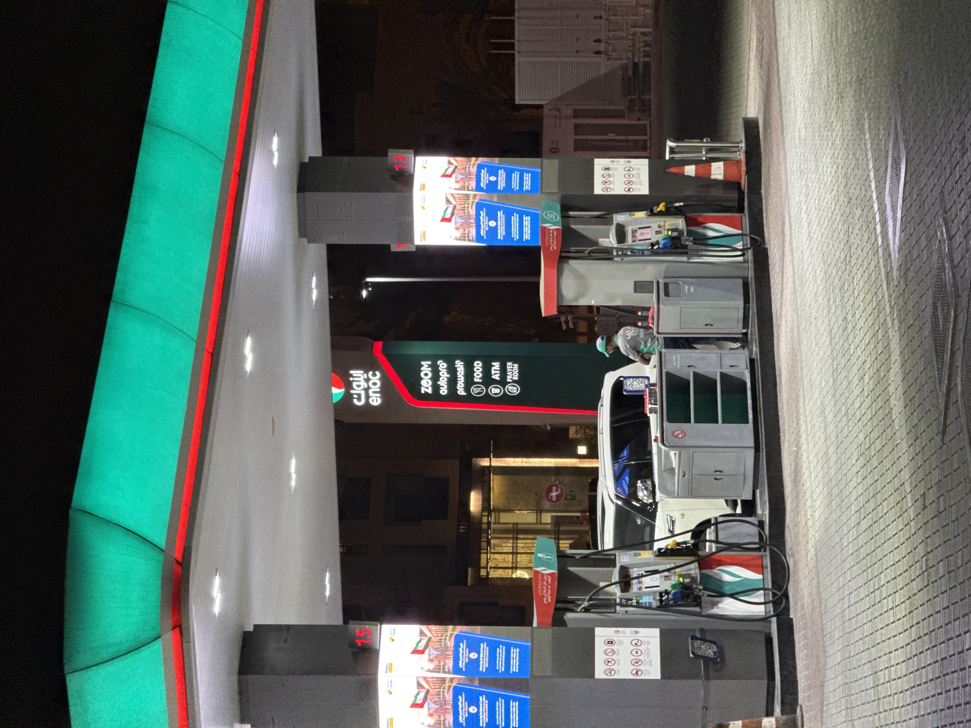 This media file captures a night scene at a gas station, likely in Dubai, United Arab Emirates, as suggested by the Arabic text on signage. The prominent subject is a white vehicle being refueled at a gas pump. A gas station attendant, wearing a green cap and a grey uniform with a logo, is visible near the pump.
The gas station itself is brightly lit, with the characteristic green and red branding of ENOC visible on a large canopy structure. Several illuminated screens display advertisements and information, including what appear to be images of the UAE flag. Various amenities are indicated on a sign, such as ZOOM convenience store, autopro car service, prowash car wash, FOOD, ATM, and PRAYER ROOM.
The foreground shows a textured pavement. The overall atmosphere is typical of a well-lit urban environment at night, with no specific weather conditions apparent. The image focuses on the transactional activity of refueling a vehicle and the services offered by the ENOC station. The visible text includes Arabic and English branding, service names, and what appears to be instructional or advertising content on the screens.