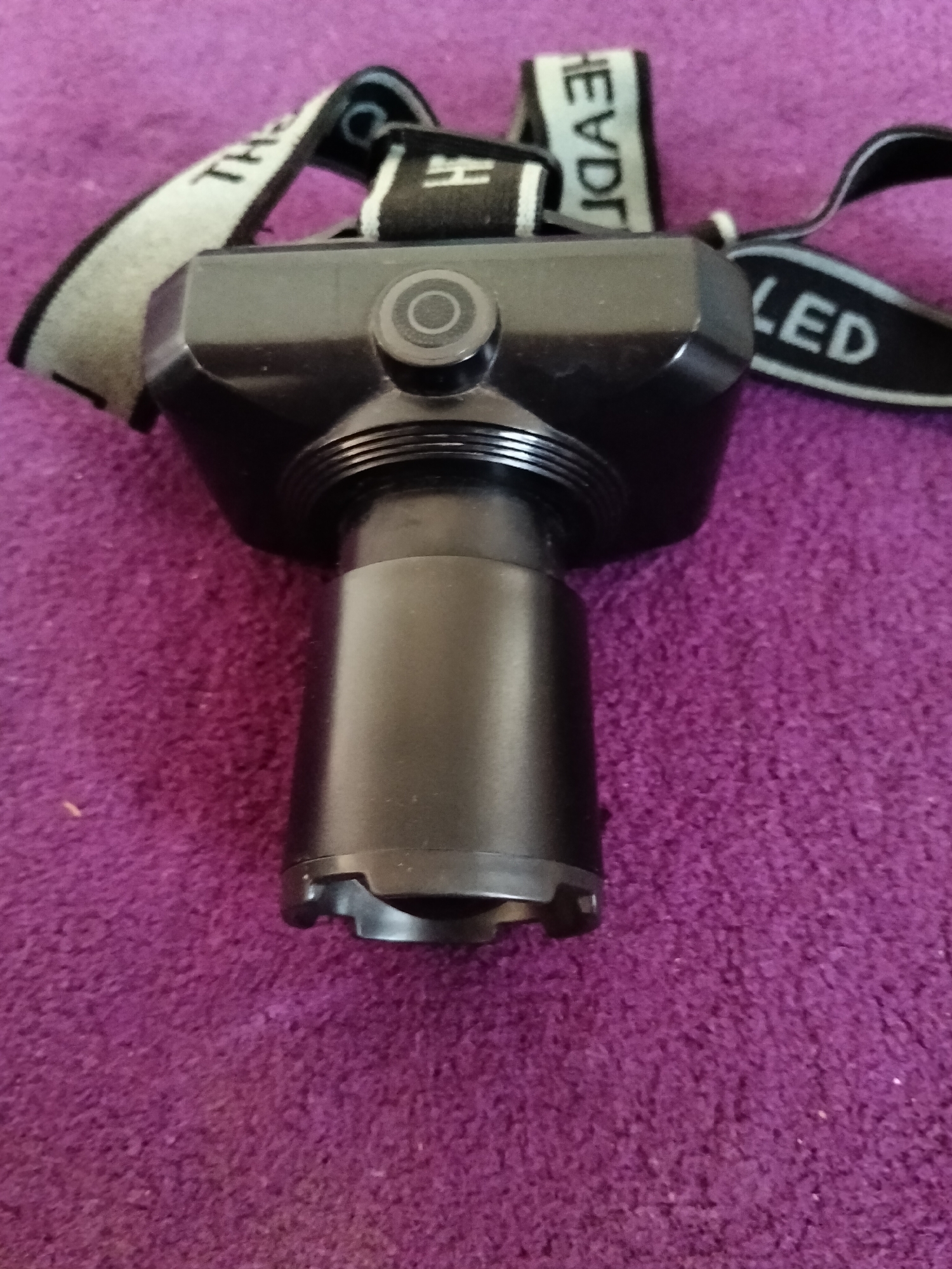 The image shows a black LED headlamp on a purple textured surface. The headlamp has a black strap with white lettering that reads "HEAD" and "LED". The headlamp itself has a cylindrical lens housing and a circular button on top. The purple surface appears to be a carpet or rug. The image is taken from a slightly elevated angle looking down at the headlamp. There are no people or any visible indicators of the location in Gempolan, Indonesia.