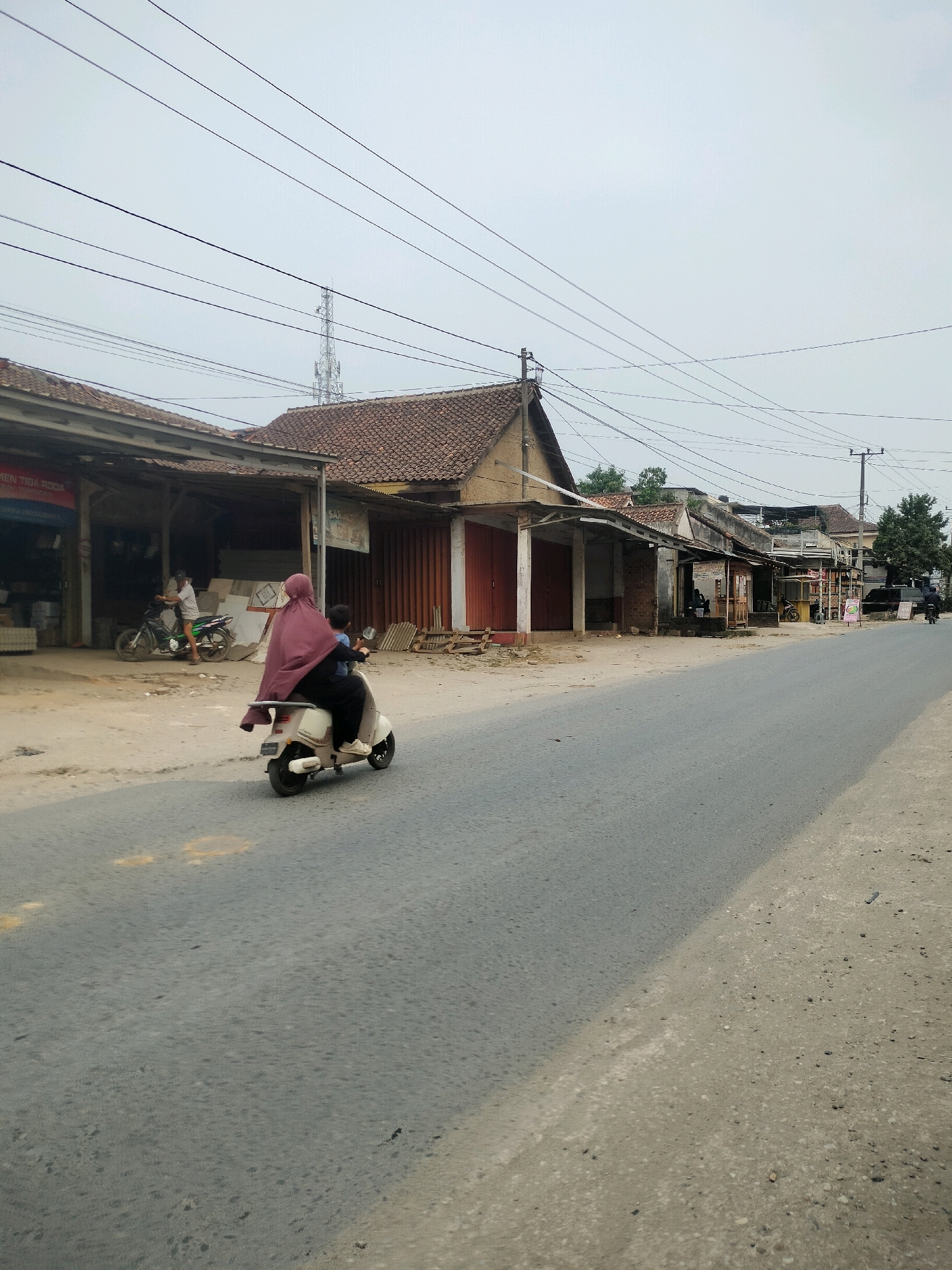 This is a photograph of a street scene in Wayakroy, Indonesia. The perspective is from eye-level, looking slightly upwards towards a hazy sky. The street is paved with asphalt and shows some wear and tear, with a sandy shoulder on the right. On the left side of the street, a row of single-story buildings with tiled roofs line the road. These appear to be shops or small businesses, some with open fronts.
In the foreground, a white scooter is moving away from the camera. The scooter is carrying two people. The rider is a person wearing a dark shirt and pants, and the passenger is wearing a maroon hijab and a dark outer garment. The passenger is seated behind the rider.
Further down the street, other elements of the environment are visible. On the left, a person on a dark motorcycle is parked in front of one of the shops. The shops have various signage, some with red lettering that is difficult to read clearly from this distance. There are also some materials, possibly building supplies, stacked near the shops.
In the background, power lines stretch across the frame, supported by wooden and metal poles. A tall telecommunications tower is visible in the distance. Further down the street, more buildings and trees can be seen. The overall atmosphere is one of a relatively quiet, rural or semi-urban street on an overcast day.