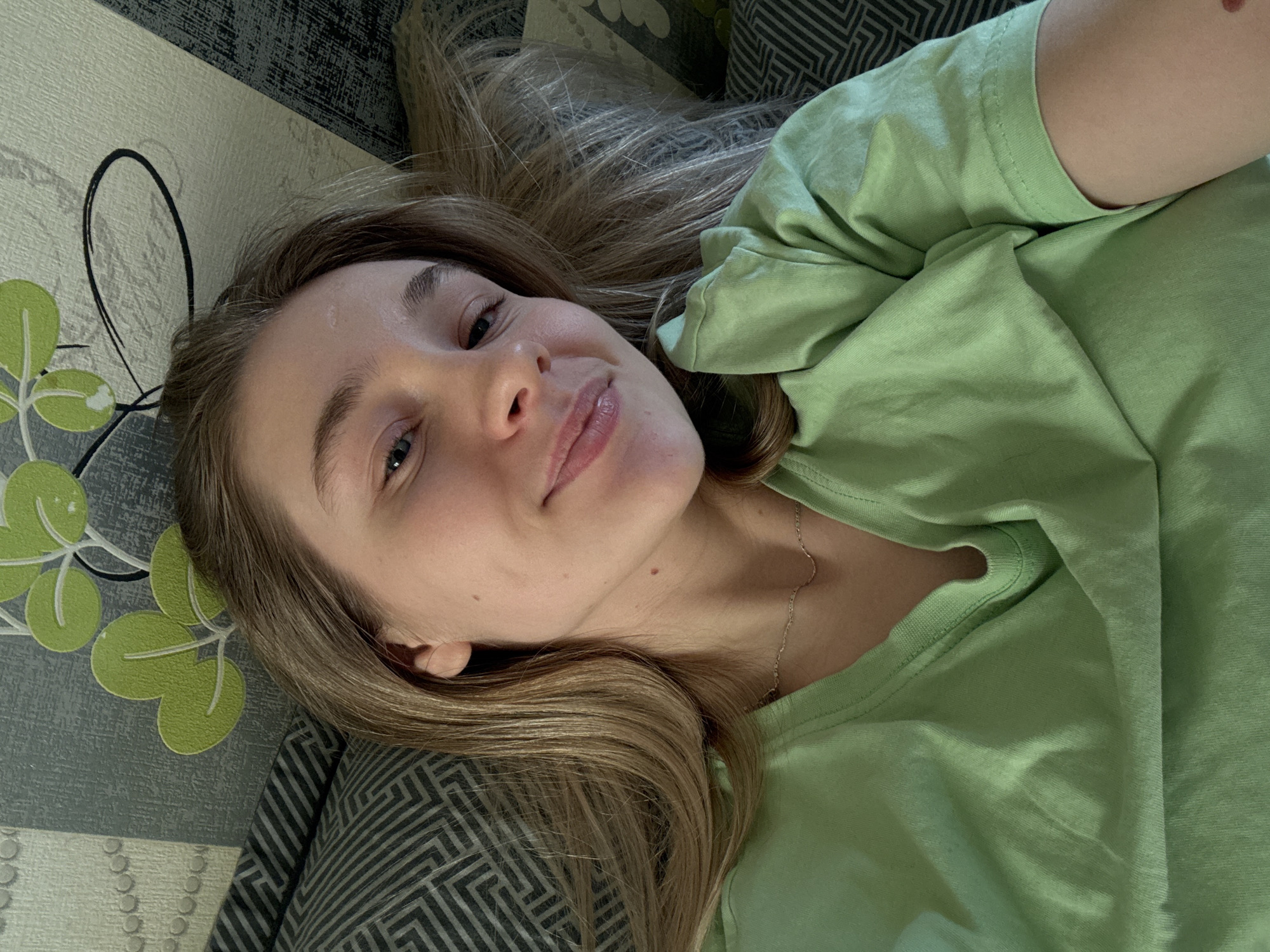 A young adult female is depicted in a close-up, head-and-shoulders self-portrait. She lies supine, looking directly at the camera with a slight smile and open eyes. Her hair is light brown/blonde, parted centrally, and extends past her shoulders. She has a light complexion, blue eyes, and visible eyelashes. A small brown mole is present near her right ear, and a small red mark is visible on her upper right arm. She is wearing a light green, short-sleeved, V-neck t-shirt and a thin, gold-colored chain necklace.
The background indicates an indoor residential setting. To her left and beneath her, patterned fabric, likely upholstery or bedding, is visible. This fabric features a dark gray base with a repeating lighter gray geometric zig-zag or Greek key pattern. A smaller section of fabric with light gray stripes and circular motifs is also present. Behind her head, the wall is covered with wallpaper featuring a light background adorned with stylized green leaves and black swirling lines. A vertical strip of plain light gray wallpaper is also visible. The lighting is even and diffused, illuminating her face without harsh shadows.
The specific location in the city of Izhevsk, Russia, cannot be determined from the visual information provided in the image.