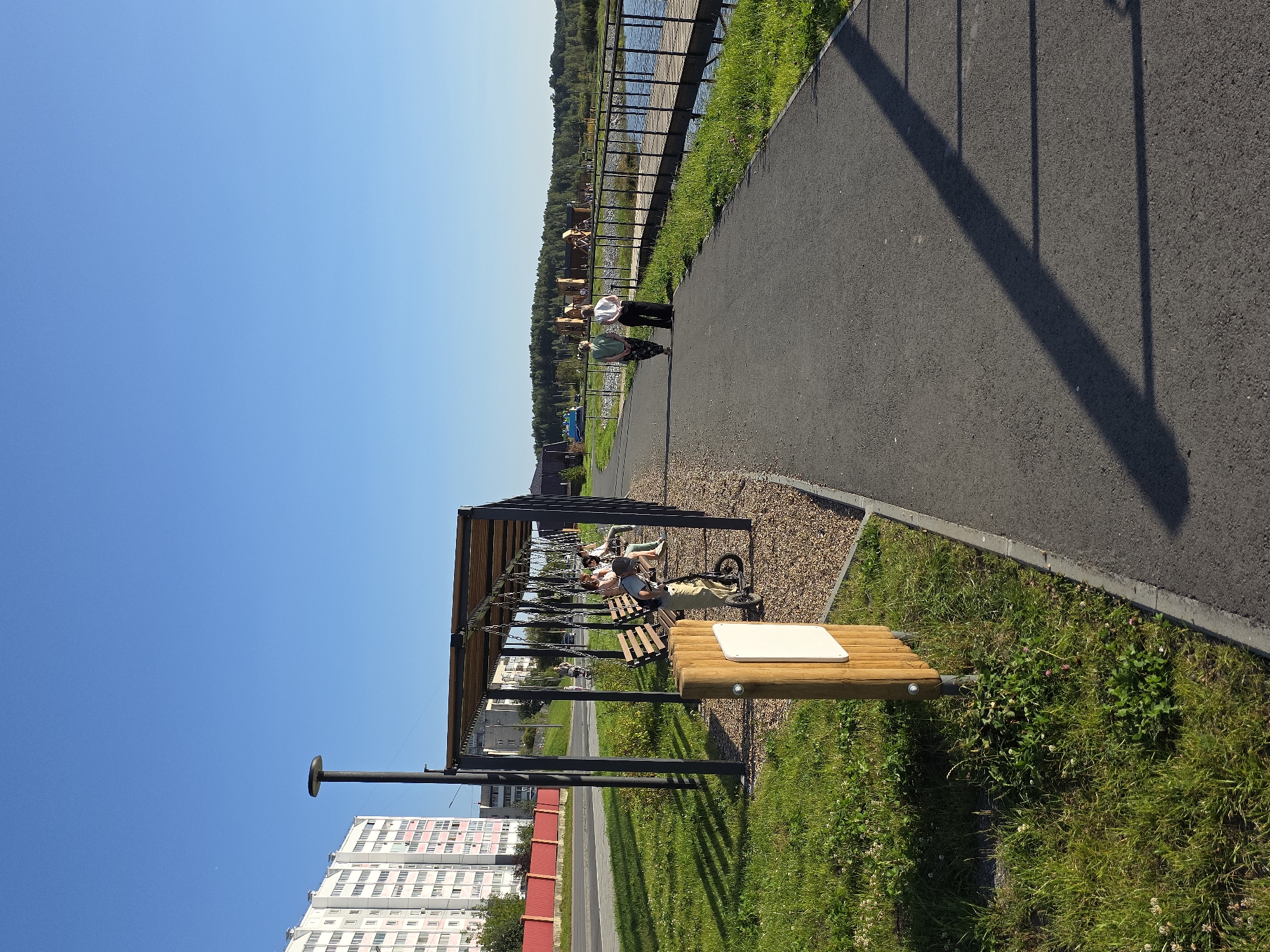 The image depicts a paved pedestrian path alongside a grassy area on the left and a body of water with a wooden boardwalk on the right. A covered seating area with swings and benches is situated to the left of the path, with several people seated on the swings and benches. Further down the path, two individuals are walking away from the camera. In the background, a multi-story building is visible, as well as trees and playground structures. The sky is clear and blue. The location appears to be a park or recreational area within the city of Miass, Russia.