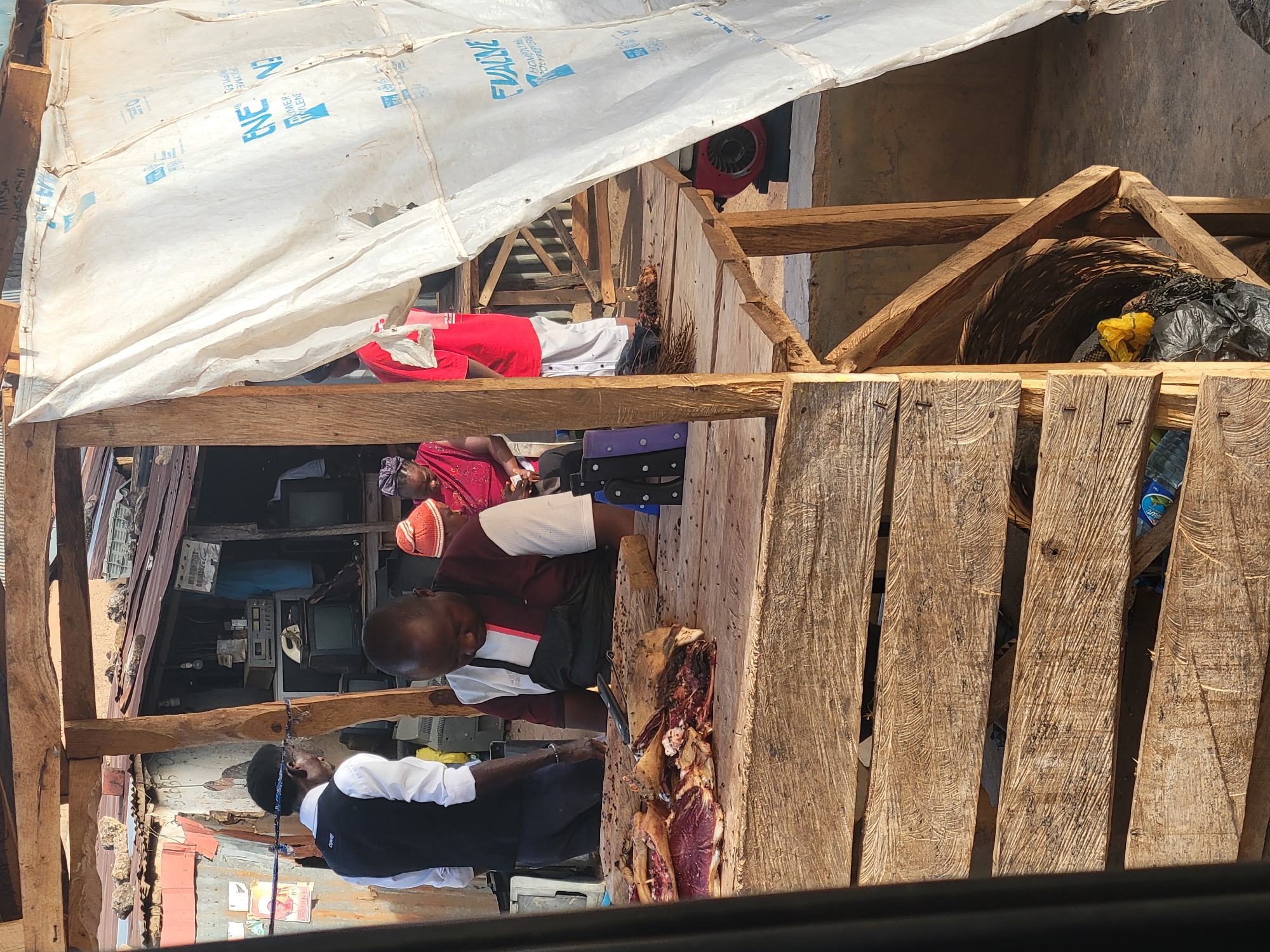 This image captures a bustling, semi-outdoor market scene in Jos, Nigeria, illuminated by bright daylight, suggesting a sunny day. The setting consists of an informal structure primarily built from rough-hewn wooden planks and posts, covered by a large, tattered white tarp serving as a canopy.
The central activity involves two men actively butchering meat on a large, thick wooden cutting surface. Substantial cuts of raw meat, likely beef or goat, are spread across the board, with visible bloodstains and several flies. One man, wearing a white shirt and black vest, is positioned on the left, focused on the task. To his right, another man in a red and white polo shirt also works at the meat stall. Behind them, a person wearing a red headscarf is partially visible, seated. Further back, another individual in red is seen lying down or resting in a shaded area.
The white tarp overhead bears blue text, partially visible and inverted in some places, showing words such as "ENE," "WATER-HYGIENE," "FOOM," and "RECYCLE," indicating it might be repurposed industrial packaging.
The background offers glimpses into other market activities and stalls. To the left, an area cluttered with old electronic devices, including several CRT televisions and what appears to be audio or repair equipment, suggests an electronics repair shop or vendor. High on the right side of the image, a red industrial fan or blower is mounted within the wooden framework. Baskets and black plastic bags, one containing yellow items, are visible on wooden shelves. Empty plastic water bottles are also scattered near the base of the wooden structure.
The scene conveys a sense of everyday commerce and labor in a rustic, vibrant market environment.