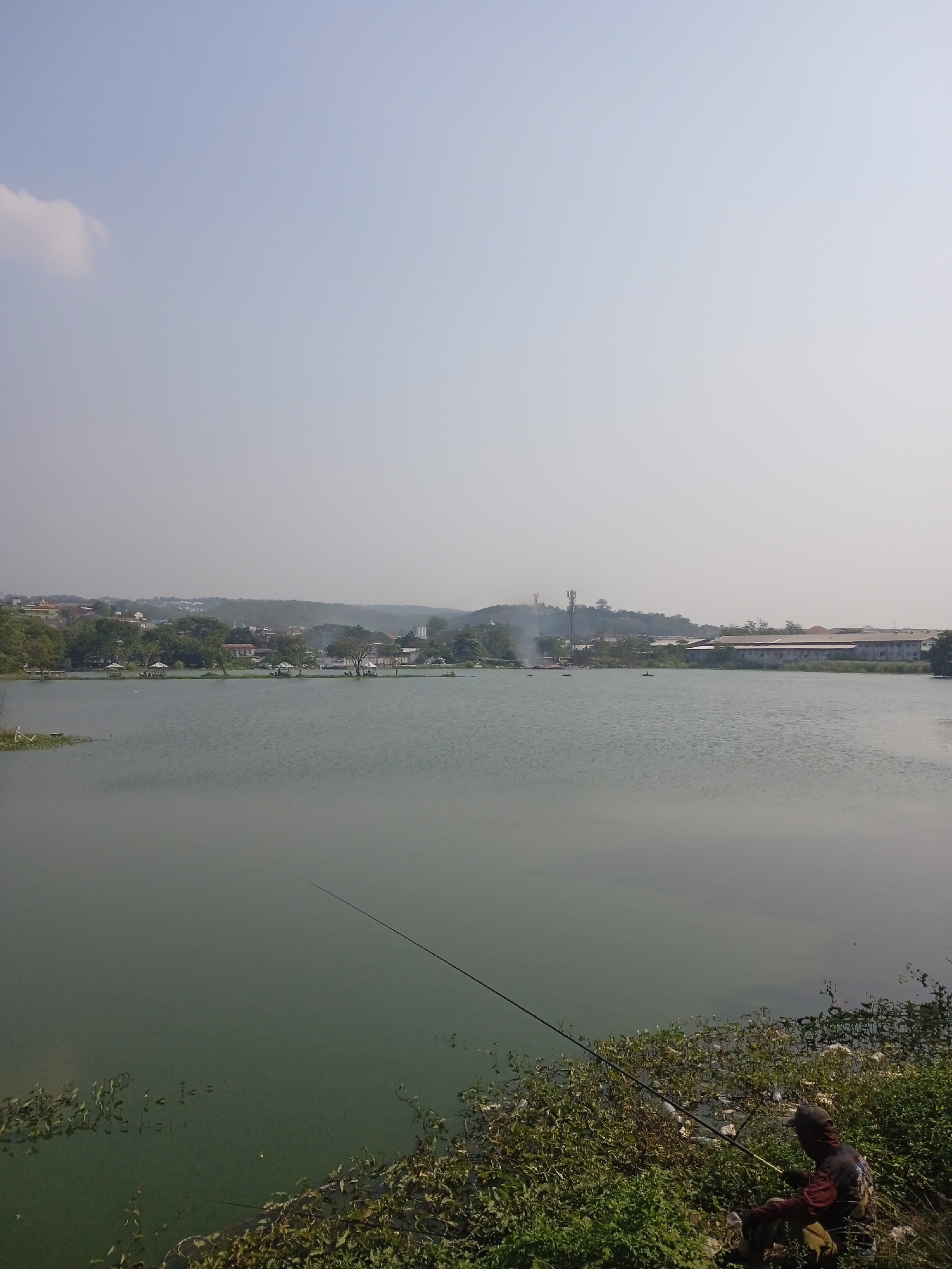 The image shows a wide, calm lake in Semarang, Indonesia. A lone fisherman sits on the grassy bank in the foreground, holding a fishing rod that extends into the water. The lake's surface reflects the hazy sky. In the background, a shoreline is visible with scattered trees, buildings, and what appear to be several small gazebos. Further back, a hillside with more buildings and vegetation rises, and a radio tower can be seen on the crest. On the far right of the background, a long, low building with a red roof is prominent, suggesting an industrial or commercial area. The overall atmosphere is peaceful and suggests a typical day in a semi-urban lakeside environment.