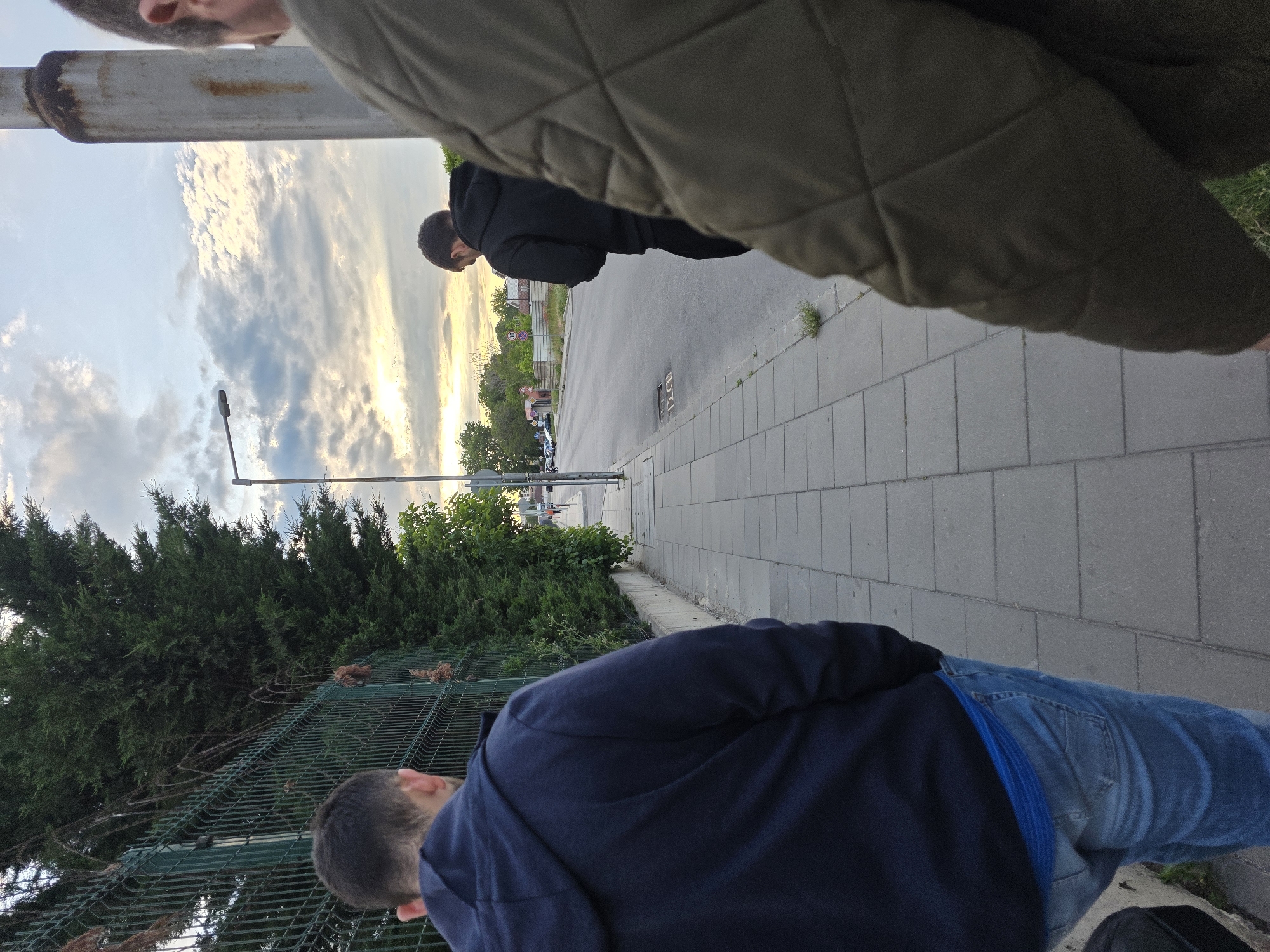 The image shows a partially obscured outdoor scene on a sidewalk in Plovdiv, Bulgaria. A person in a navy blue hoodie and blue jeans is positioned in the foreground, facing away from the camera, near a green metal fence. Another person wearing a black hoodie stands further down the sidewalk, partially obscured by someone wearing a quilted olive-green jacket closer to the camera. The sidewalk is paved with gray rectangular slabs and borders a street. A lamppost is visible. Across the street are buildings and trees, with a bright sky and some clouds. The image is taken at a low angle.