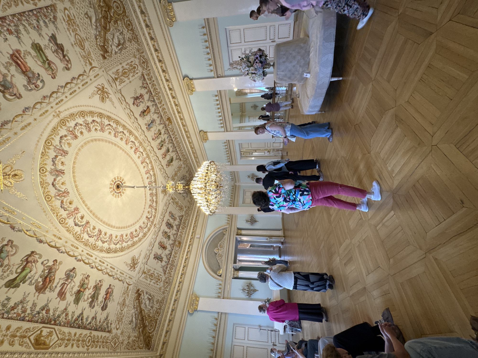 The image captures a group of people in a grand, ornate ballroom in Saint Petersburg, Russia. The high ceiling is intricately decorated with gilded stucco work and painted frescoes depicting mythological figures. A large, elaborate chandelier hangs from the center of the ceiling, illuminating the room. The walls are painted a pale blue and adorned with classical architectural details and decorative moldings. The floor is made of polished wooden parquet in a geometric pattern.
The people in the image appear to be a tour group or visitors, standing and observing the architecture or an exhibit. Some individuals are looking up at the ceiling, while others are focused on something further into the room, possibly a display or another architectural feature. The attire of the people suggests a daytime visit, and the atmosphere seems to be one of quiet observation and appreciation for the historical setting. One person in the foreground is wearing a brightly colored, patterned shirt and pink pants, and another is in a white sweater with a black and white patterned skirt. The overall impression is one of a cultural visit to a significant historical landmark.