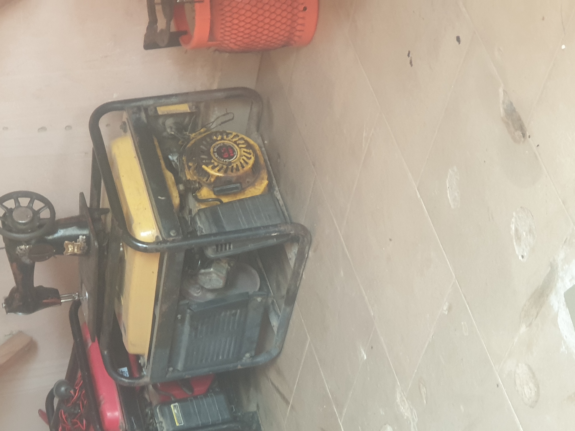 In Jauro Yinu, Nigeria, an indoor scene shows a yellow and black generator with a sewing machine on top. To the left of the generator is a red object, possibly another generator. To the right, an orange mesh basket is visible. The objects are placed on a tiled floor. The lighting suggests an indoor setting, potentially a storage area or workshop.