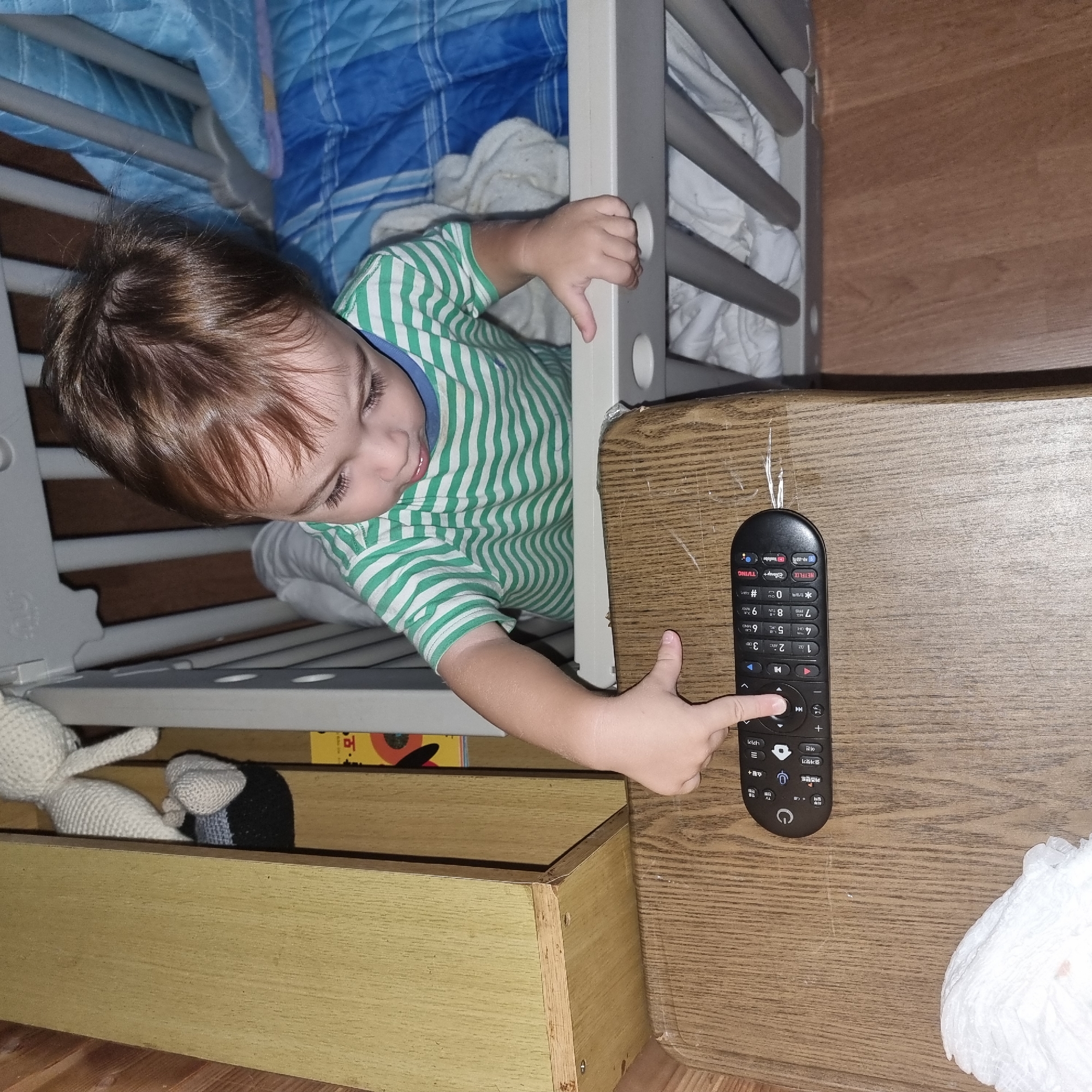 A toddler, wearing a green and white striped shirt, is positioned inside a crib, leaning over the edge and reaching for a black remote control on a wooden table. The remote has several buttons, including a power symbol, volume controls, number keys, and streaming service icons. The crib contains white bedding and is adjacent to a wooden shelf unit. The floor is wooden. The scene is lit by artificial light. The image is a close-up, focusing on the toddler's interaction with the remote and the immediate surroundings.