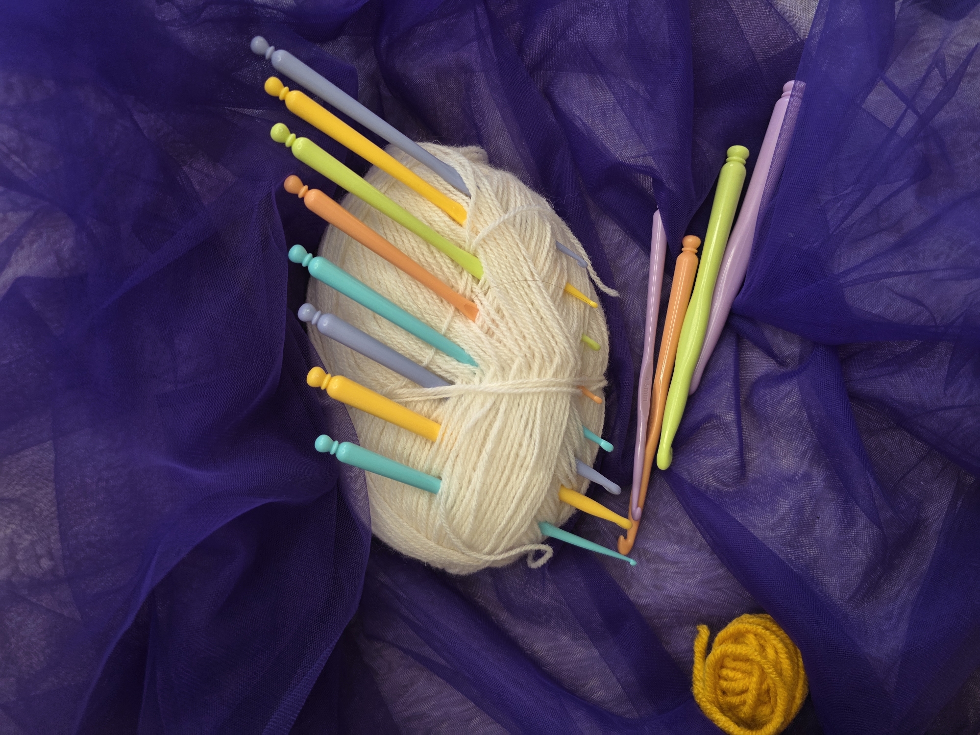 The image shows a ball of off-white yarn with several colorful crochet hooks stuck into it. There is also a small ball of yellow yarn. All of this is set against a draped background of purple sheer fabric. The crochet hooks are various colors, including purple, green, yellow, orange, and turquoise. The location is unknown, and it's also unknown if this image was taken in the city of Chaikovsky, Russia.