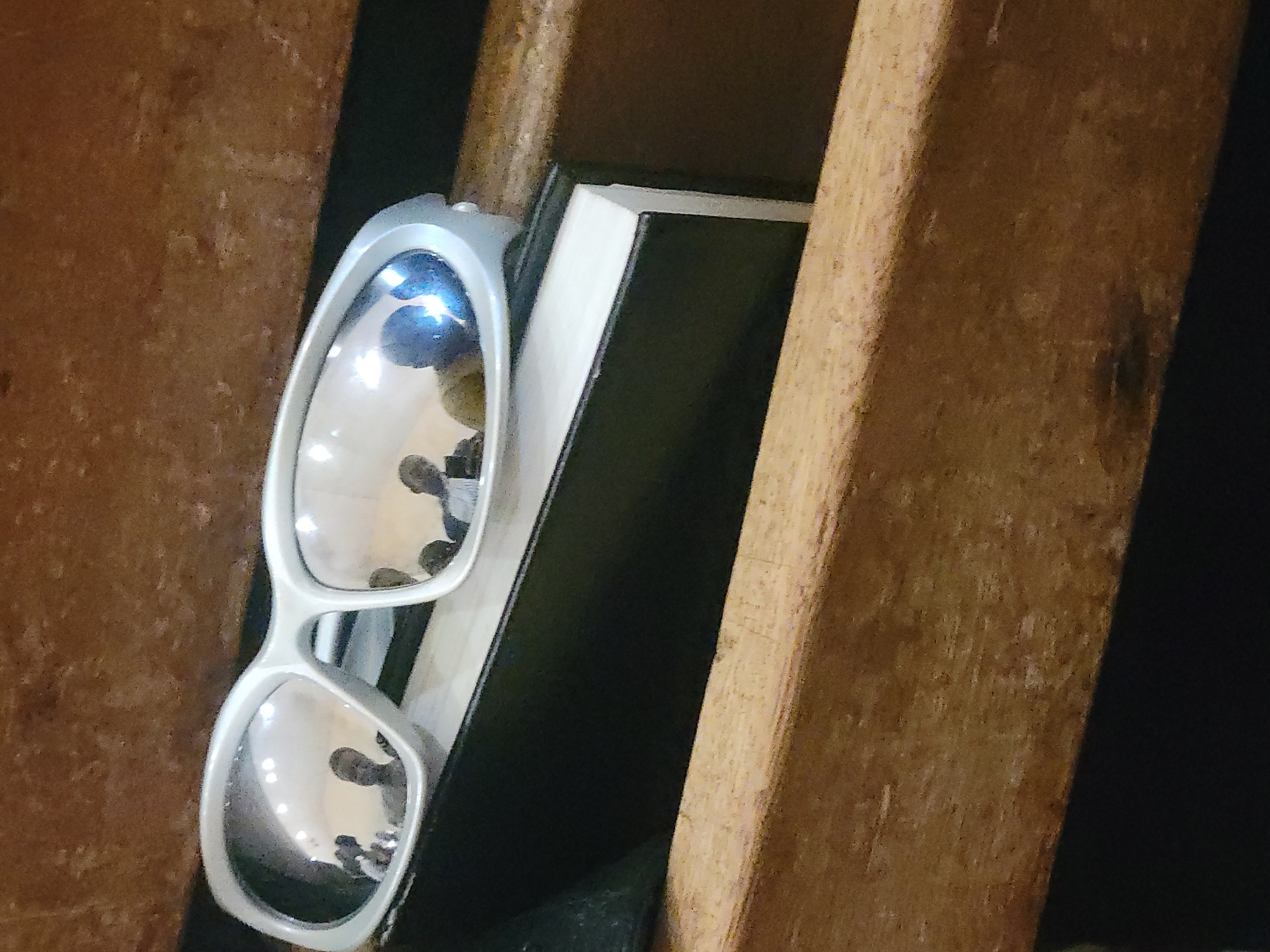 A close-up shot shows a pair of white-framed sunglasses resting on a book. The glasses are positioned between two wooden beams. The scene appears to be indoors, and the book is dark-colored with white pages. The location is not discernible from the image.