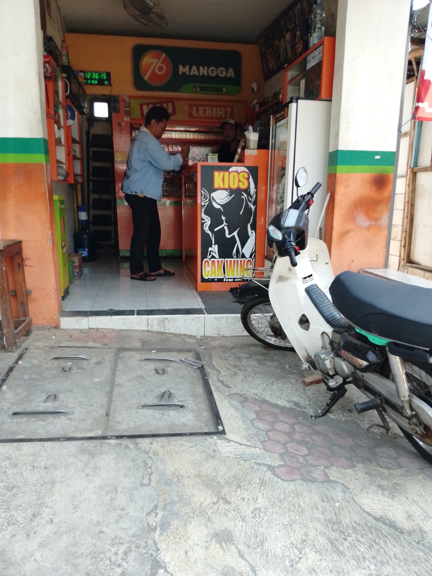 A person wearing a blue denim jacket and black pants stands behind a counter in a small shop. The shop appears to be an outdoor kiosk with orange and green painted walls. A digital clock on the wall displays "12:35:15". A white motorcycle is parked in front of the shop. The ground in front of the shop is made of concrete and has two metal manhole covers. The scene is likely located in a street or public area in Pasuruan, Indonesia, given the text on the signs, which appears to be in Indonesian.