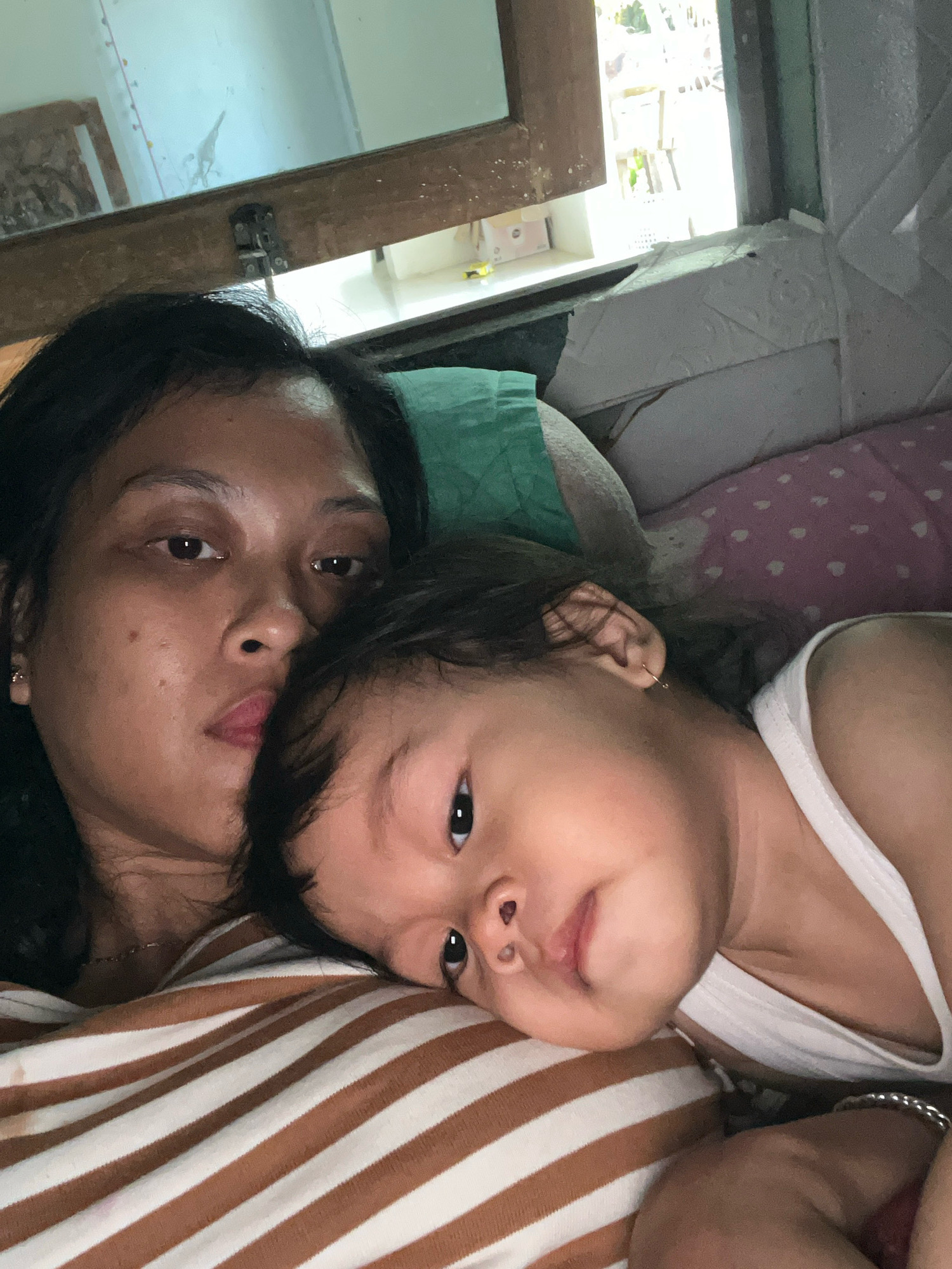 In Kecamatan Pati, Indonesia, a close-up, low-angle shot shows a woman and a baby lying on a bed. The woman, positioned on the left, has dark hair and is looking towards the camera with a neutral expression. Her right ear, adorned with a small hoop earring, is visible. Beside her, the baby, on the right, rests its head on the woman's chest, with its body angled away from the camera. The baby has dark hair and is looking upwards, its eyes wide. The scene is lit by natural light coming from a window in the background. The bed is covered with striped bedding in brown and white, and there are pillows in pink with white polka dots and green accents. The environment suggests a domestic setting, likely a bedroom.