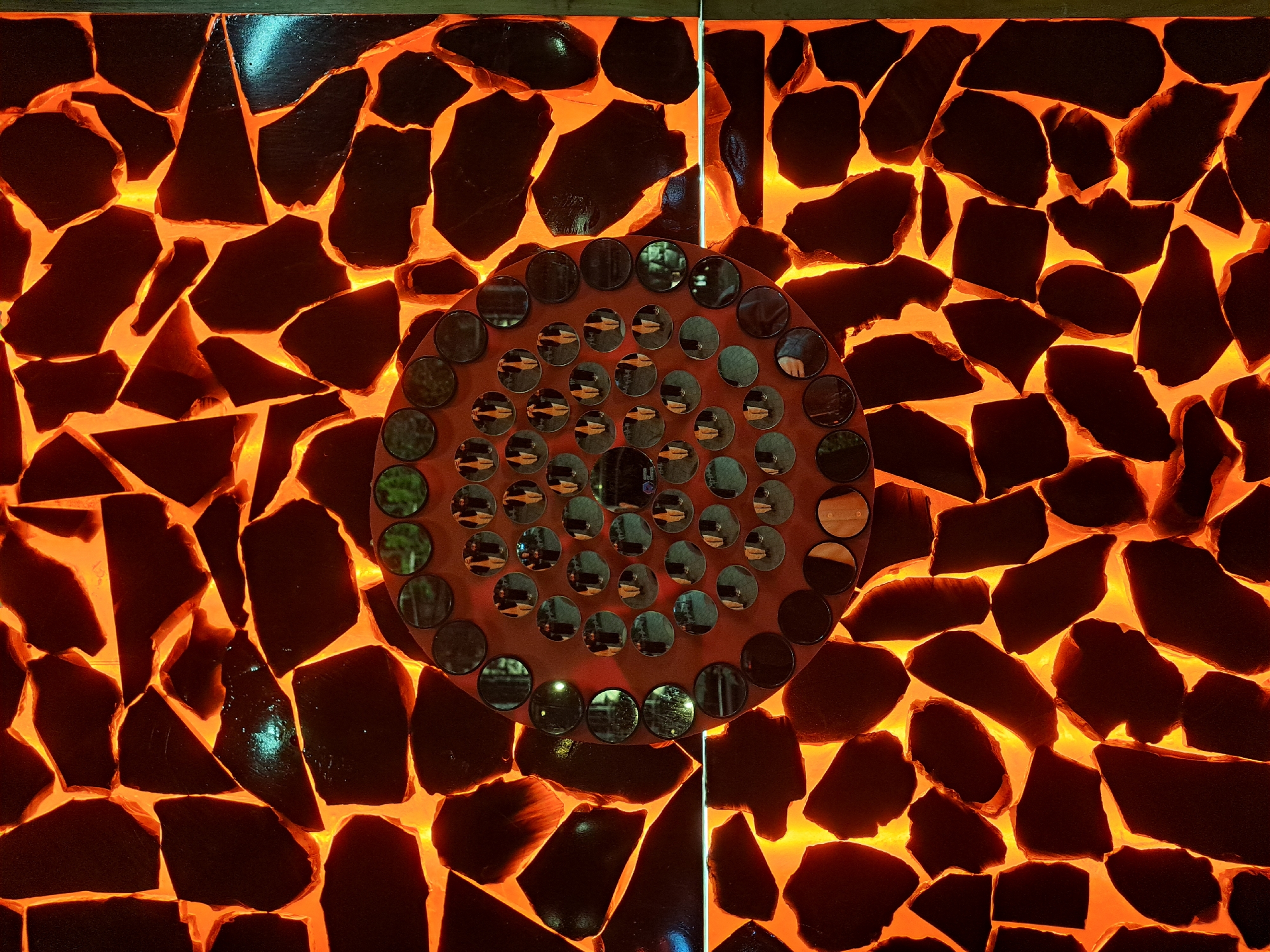 The image depicts a wall decoration in Yerevan, Armenia. The wall is constructed from dark, irregularly shaped stones or tiles, with bright orange light emanating from the spaces between them, giving the impression of molten lava. A circular decorative element is affixed to the wall. This circular element contains many small, round, mirror-like discs that reflect the surrounding environment and people. The wall appears to be part of an interior design scheme, with a modern and potentially artistic aesthetic. The lighting and design create a warm and visually striking display.