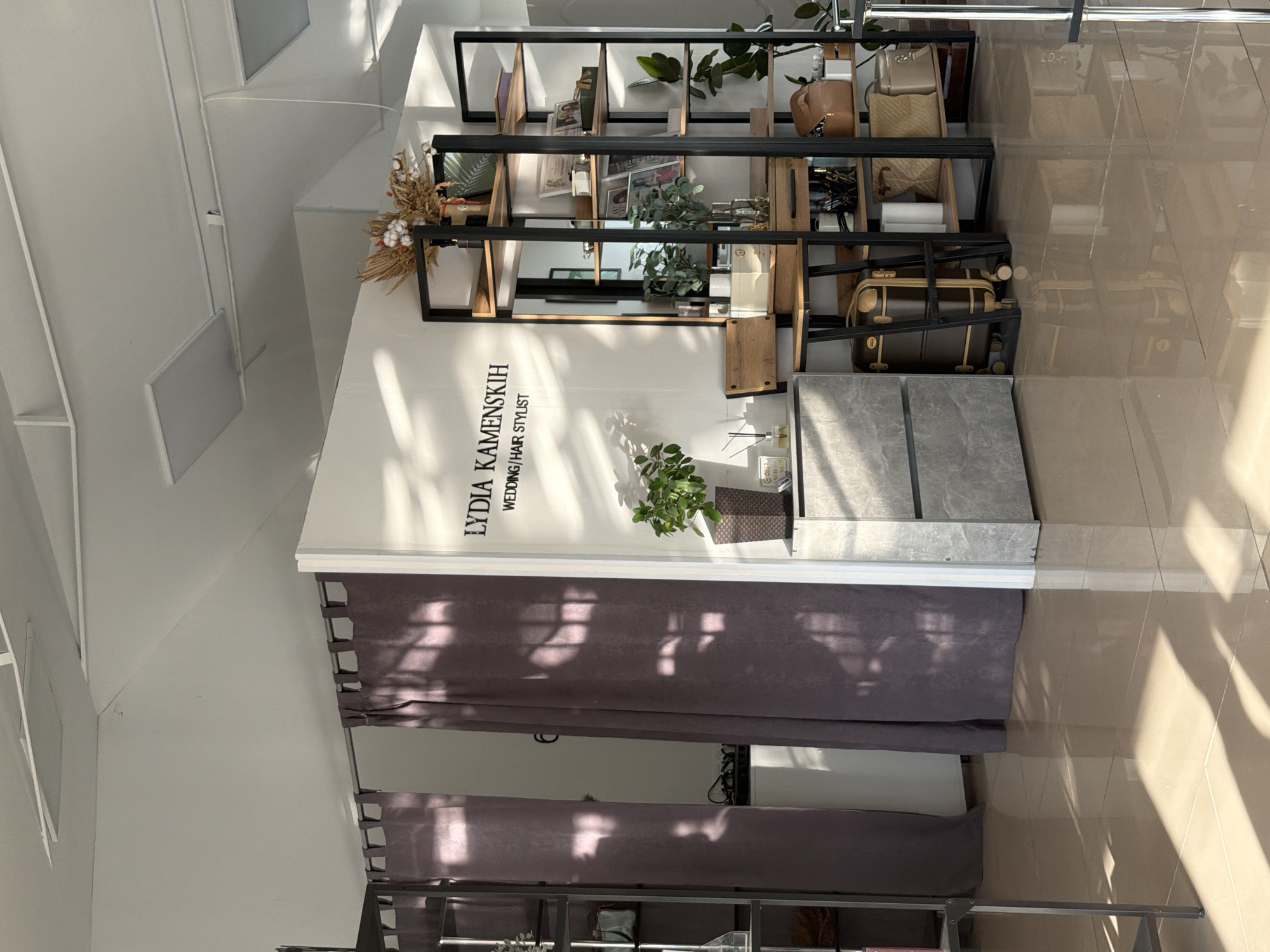 The image displays the interior of a professional establishment, identified by signage as "Lydia Kamenskikh, Wedding/Hair Stylist," located in Chaikovsky, Russia. The space is brightly illuminated by natural sunlight, casting distinct, angular shadows across the light-colored, reflective floor and dark gray curtains, indicating a daytime setting.
The white ceiling features recessed lighting or vents. Below, a prominent black metal and wood shelving unit displays various decorative items such as small potted plants, dried floral arrangements, books, framed pictures, handbags, woven baskets, and a vintage-style brown suitcase on the lowest shelf. The aforementioned black sign with white text is mounted on the white wall behind the shelving unit.
To the right and below the sign, a small green plant in a pot rests on a low surface, adjacent to a gray, textured block-like structure that serves as a display stand or counter. Dark gray curtains, which appear to have horizontal pleats or folds, are partially drawn across a section of the room, marked by the strong sunlight creating patterned shadows on their surface. The overall ambiance is clean, organized, and professionally decorated, suggesting a welcoming commercial space. No people are visible in the scene, and no specific activity is taking place.