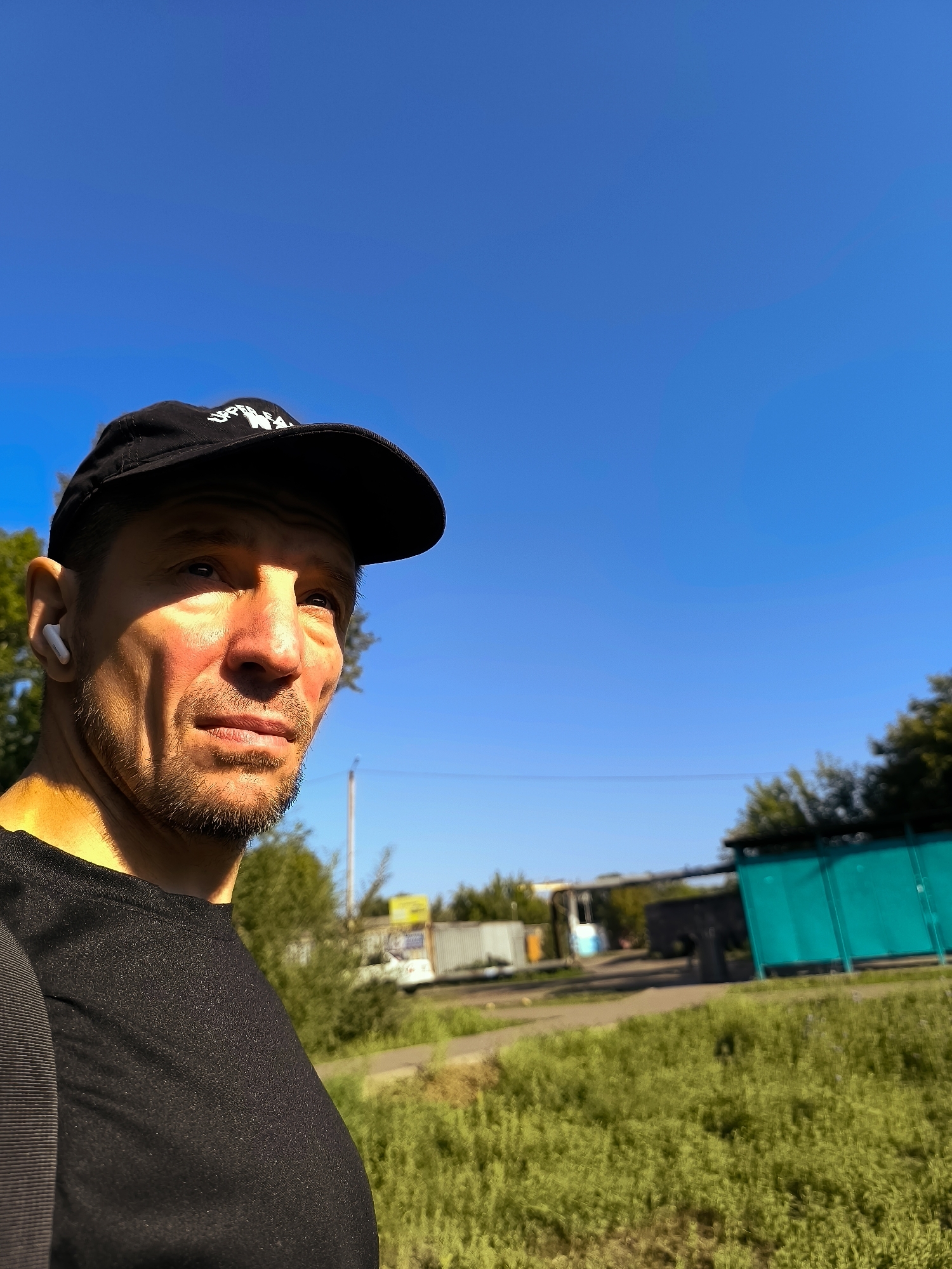 A man wearing a black t-shirt and a black baseball cap with white lettering stands in the foreground, looking to his right. He has a light beard and is wearing white wireless earbuds. The background consists of a bright blue sky, green grass, trees, and a turquoise fence. There are some buildings and structures visible in the distance, suggesting an urban or semi-urban environment. The lighting indicates it is daytime with the sun casting shadows on the man's face. The overall scene appears to be outdoors.