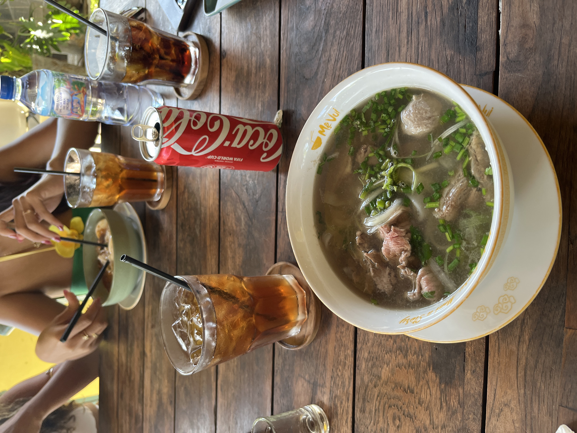On a wooden table at a restaurant in North Kuta, Bali, Indonesia, a bowl of Vietnamese pho noodle soup is presented. The soup contains thinly sliced beef, meatballs, white noodles, and green onions. A can of Coca-Cola and three glasses of iced tea are also on the table. In the background, two women are visible, one is stirring the soup with chopsticks while the other is holding a flower. The restaurant appears to be an outdoor seating area with tropical plants visible.
