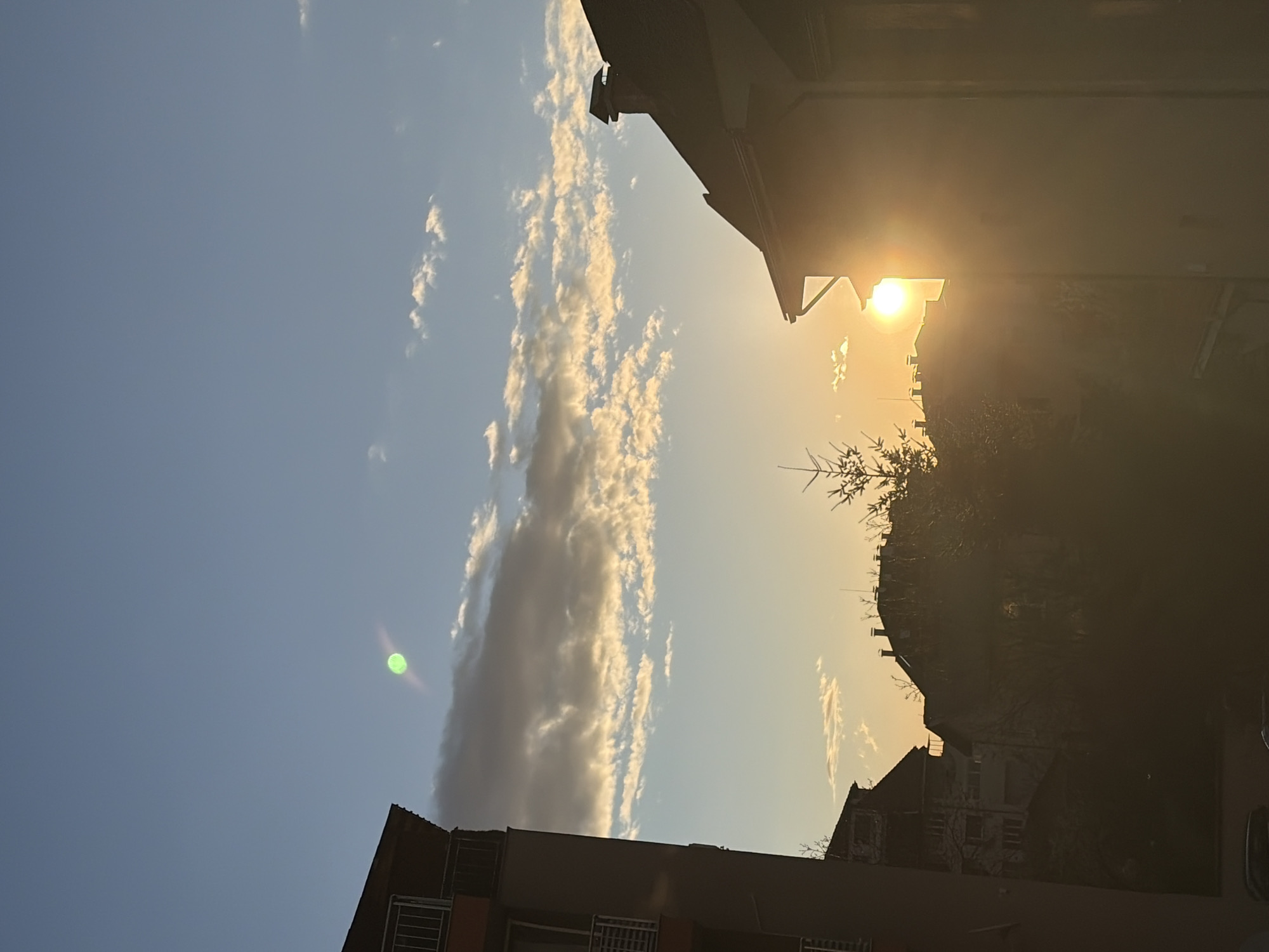 The image captures an outdoor scene, likely in the city of Sofia, Bulgaria, during a sunset or sunrise. The sun is visible, partially obscured by buildings, creating a bright glare. Buildings are visible in the foreground and background, with the sky featuring elongated clouds. The image appears to be taken from a low angle. The buildings are multi-story structures, possibly residential or commercial. There are also some trees in the scene.